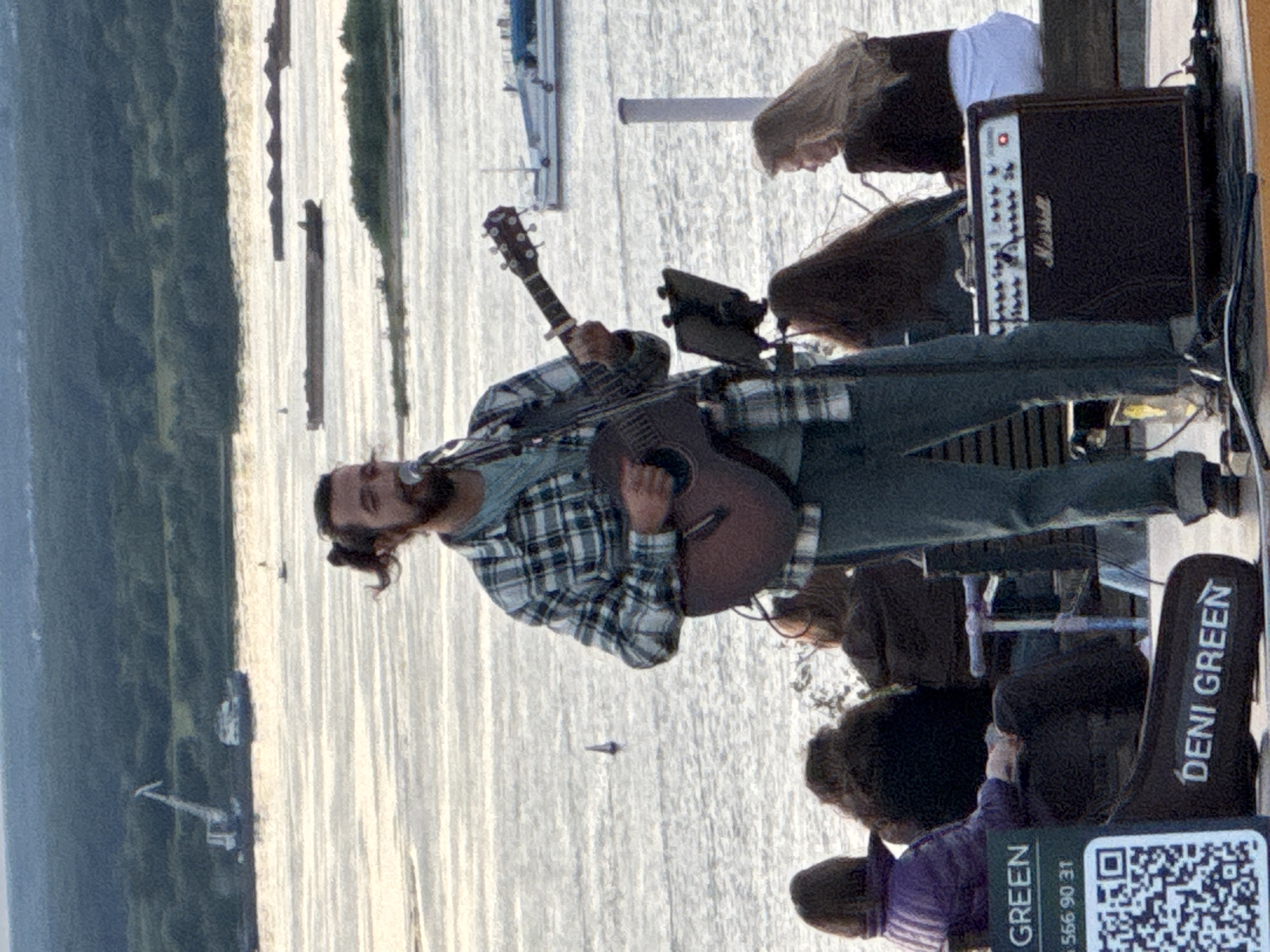 The image depicts an outdoor concert or performance by a male musician playing an acoustic guitar and singing into a microphone. He has a man bun and is wearing a plaid shirt and jeans. In the foreground, an amplifier with the "Marshall" logo is visible. There are several audience members seated behind the performer, facing away from the camera. To the left, a sign with "DENI GREEN" and a QR code is present. The background shows a wide body of water, likely the Volga River, with several boats and barges visible, as well as a distant treeline and industrial cranes. The setting appears to be a waterfront area in Nizhny Novgorod, Russia, possibly at a park or embankment, given the presence of public seating and the general ambiance of a riverside event. The lighting suggests it is either early morning or late afternoon.
