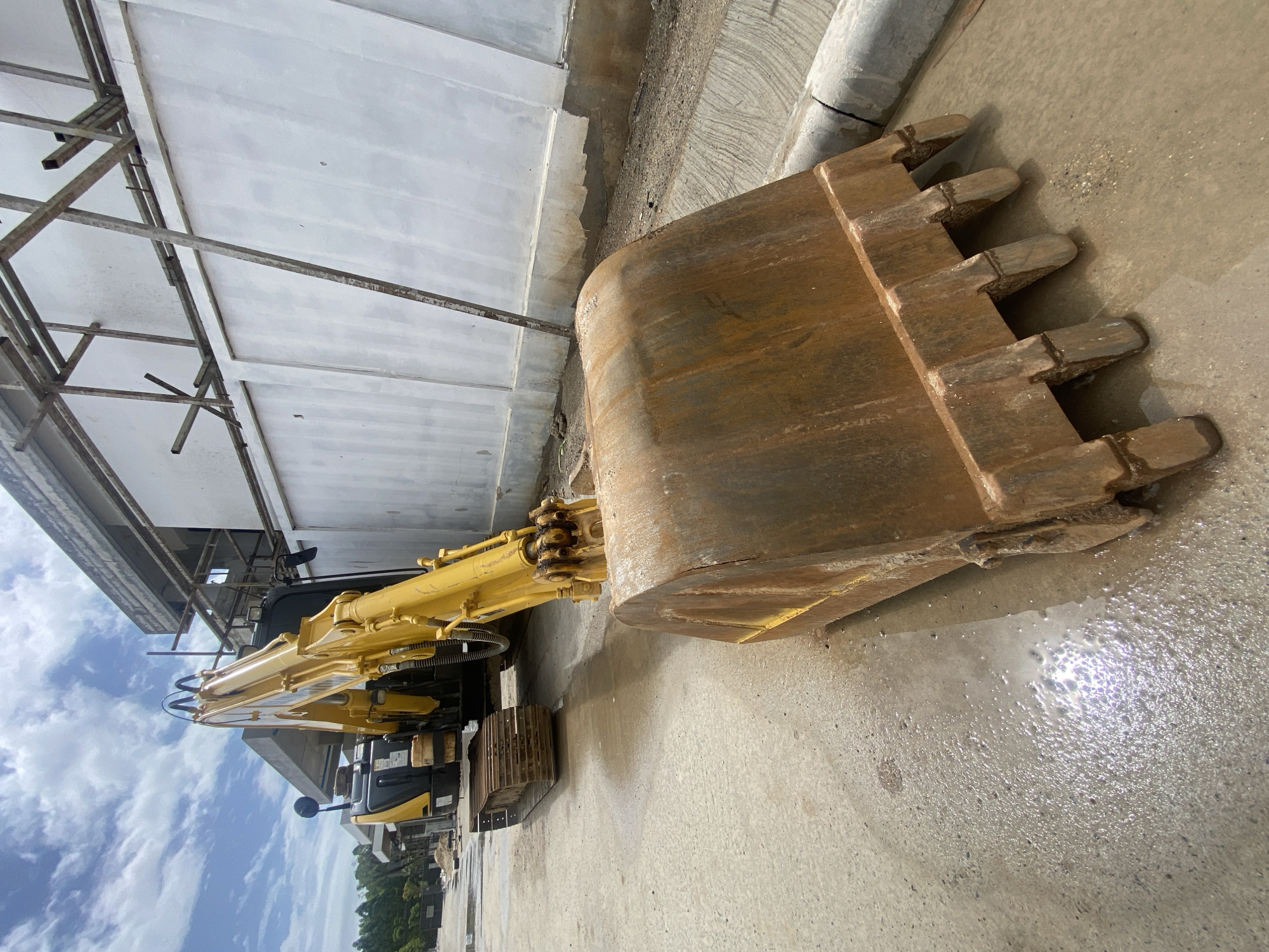 A yellow Komatsu excavator is parked on a concrete surface next to a white building with metal framework, in Choeng Thale, Thailand. The excavator's bucket faces the viewer, showing its worn condition. The ground is wet, reflecting the sky. The excavator is positioned parallel to the building. In the background, trees and a cloudy sky are visible.