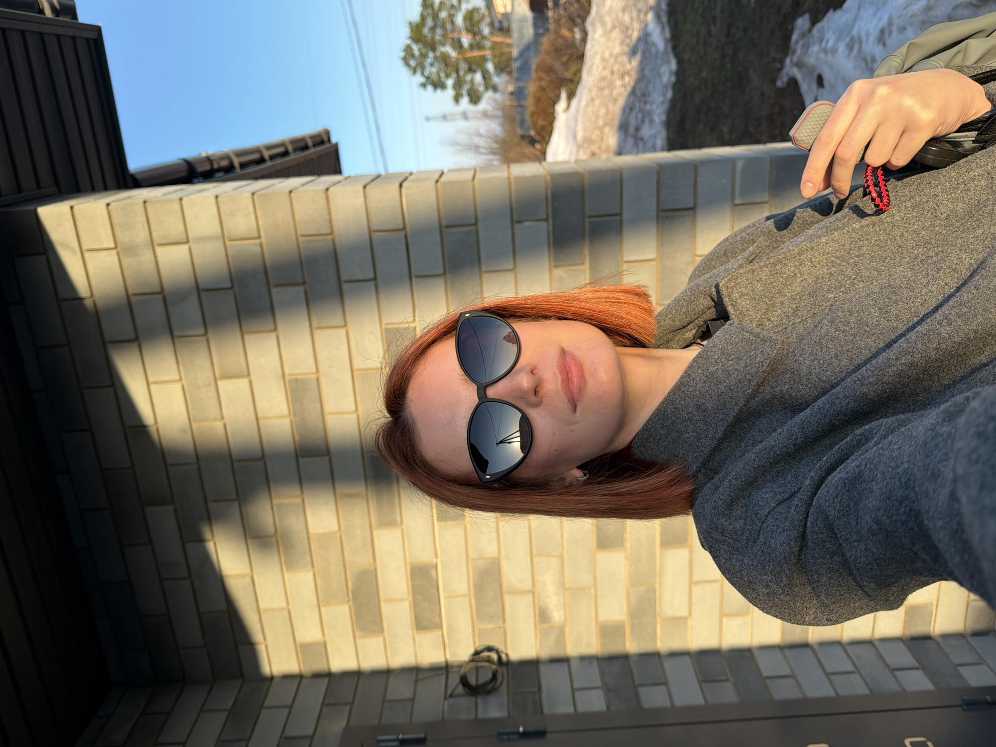 A woman with shoulder-length auburn hair, wearing dark sunglasses and a gray hoodie, poses for a photograph outdoors. She stands in front of a light-colored brick wall, partly in shadow. Behind the wall, there is a patch of melting snow and a tree. The image's location is Chaikovsky, Russia. The photo is slightly rotated clockwise. The woman is holding an object with a red and black cord in her right hand. The location seems to be a private residence due to the brick wall and door in the foreground. The sky is clear blue.