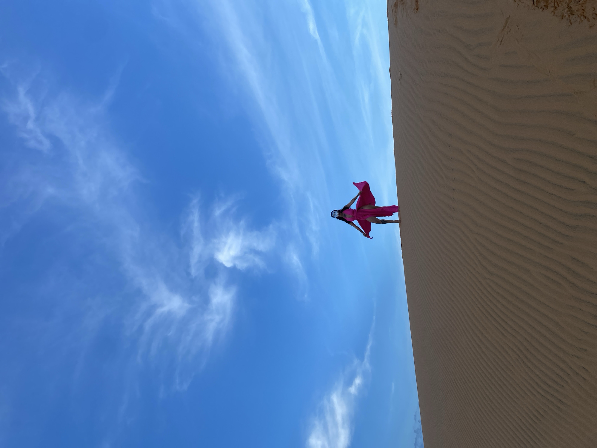 A lone figure stands atop a sand dune in Mũi Né, Vietnam, against a bright blue sky with wispy white clouds. The person is wearing a long, flowing pink dress and a mask. The dress billows out around them, creating a dramatic silhouette. The sand dune is characterized by rippled patterns created by wind. The image captures a serene and visually striking moment in the desert landscape.