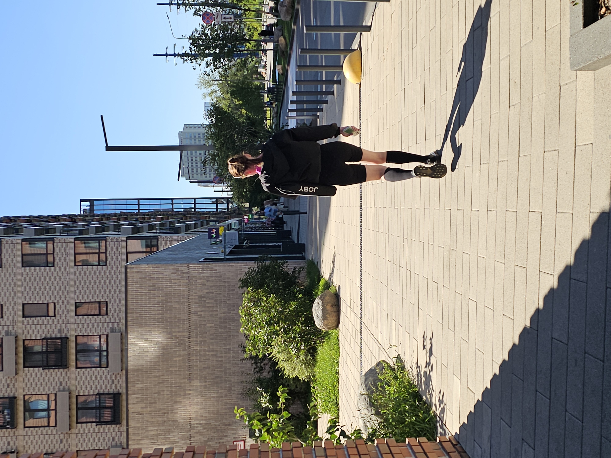 In a modern urban setting in Moscow, Russia, a woman is walking away from the camera on a paved pedestrian walkway. She is wearing athletic attire: black shorts, black compression socks, and a black jacket. A black bag with the "JOBY" logo is slung over her right shoulder. In her left hand, she holds a light-colored object, possibly a water bottle. The walkway is lined with bollards and a yellow speed bump is visible on the right side. To the left of the walkway, there is a grassy area with large stones and plants, adjacent to a brick wall and a multi-story building with many windows and balconies. In the background, there are more buildings, trees, streetlights, and power lines, indicating a street with moderate traffic. The sky is clear blue, suggesting a sunny day. The shadows cast by the woman and other objects indicate the sun is positioned to the left and slightly behind the viewer.