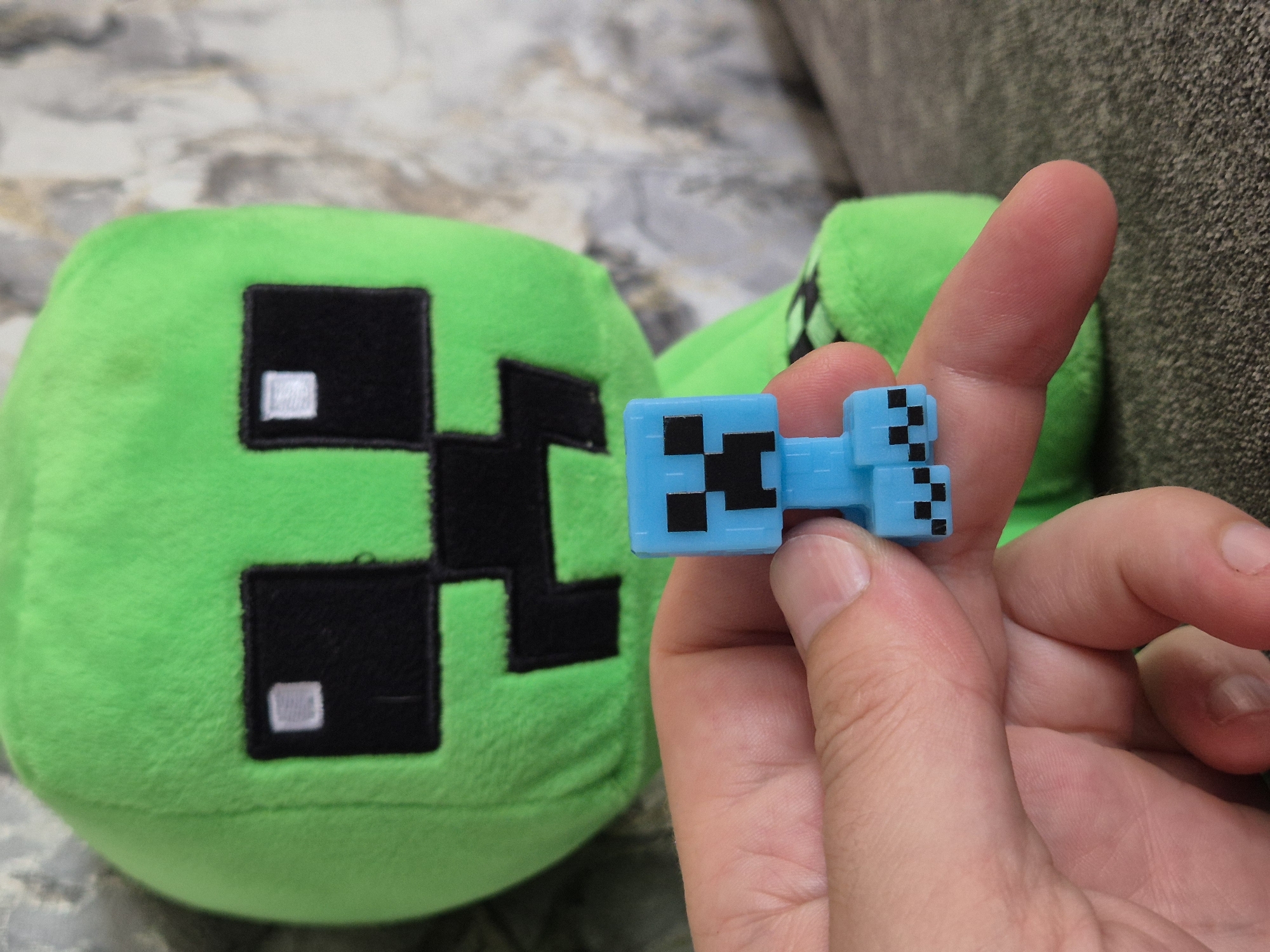 A person's hand holds a small, blue, pixelated figurine of a Minecraft character. The figurine is held between the thumb and index finger, with a portion of the index finger visible in the foreground. In the background, a large, plush green toy resembling a Minecraft Creeper is visible, with its characteristic black square eyes and mouth. The setting appears to be indoors, with a textured surface, possibly a couch or upholstered furniture, visible below the hand. The background behind the plush toy has a mottled, light-colored texture, perhaps a wall or curtain. There is no indication of the city of Taldykorgan, Kazakhstan, in this image.