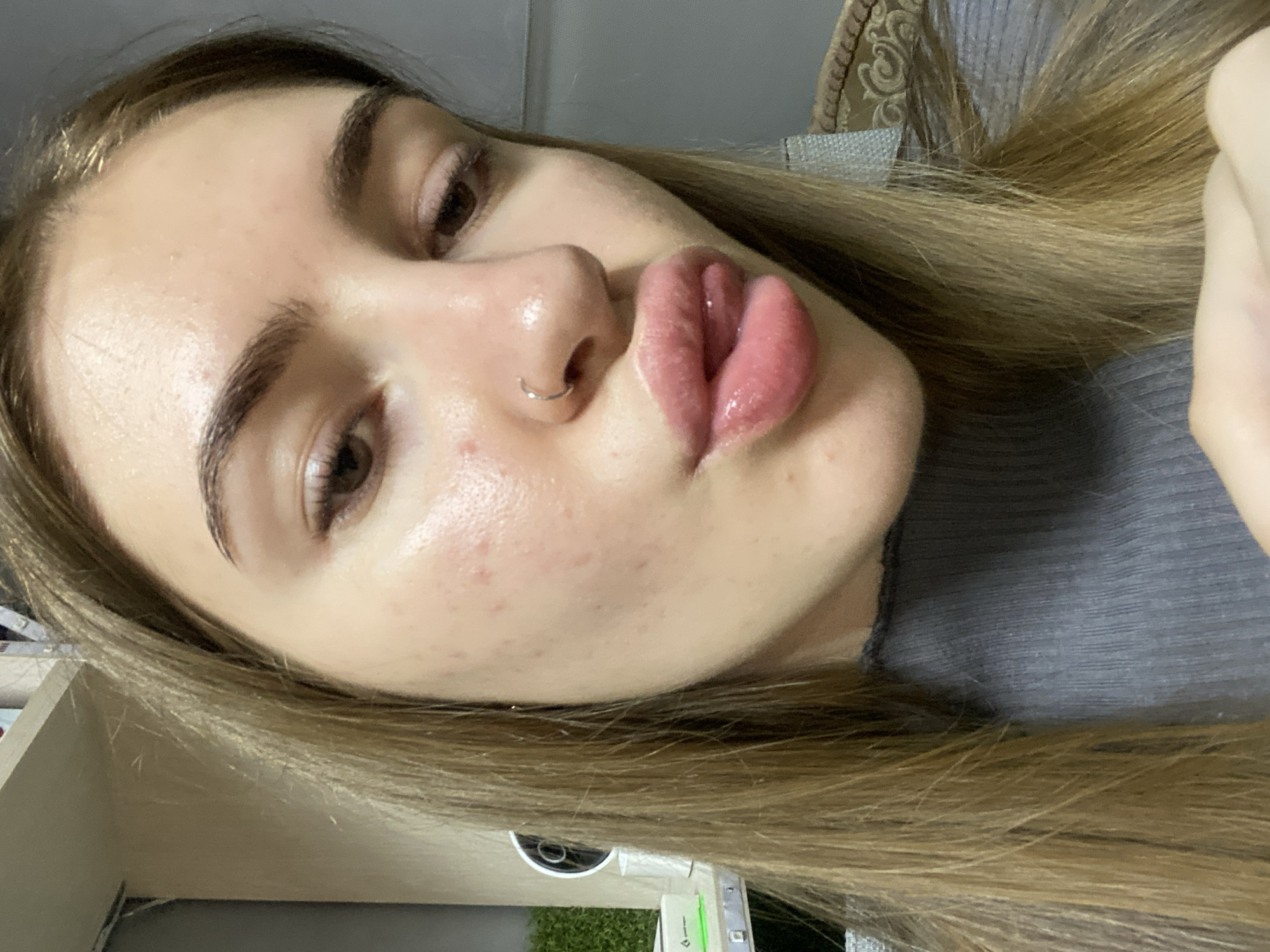 A medium shot, close-up selfie of a young woman with blonde hair is presented. She is looking downwards and to the right, with her lips pursed and her tongue slightly extended. The woman is wearing a gray ribbed turtleneck. She has a silver hoop nose ring. Her skin appears to have some texture and redness, particularly on her cheeks. The background is blurred, but suggests an indoor environment with shelves and possibly a plant on the left side. No specific details about the location in the city of Дьяково, Russia are discernible from the image.