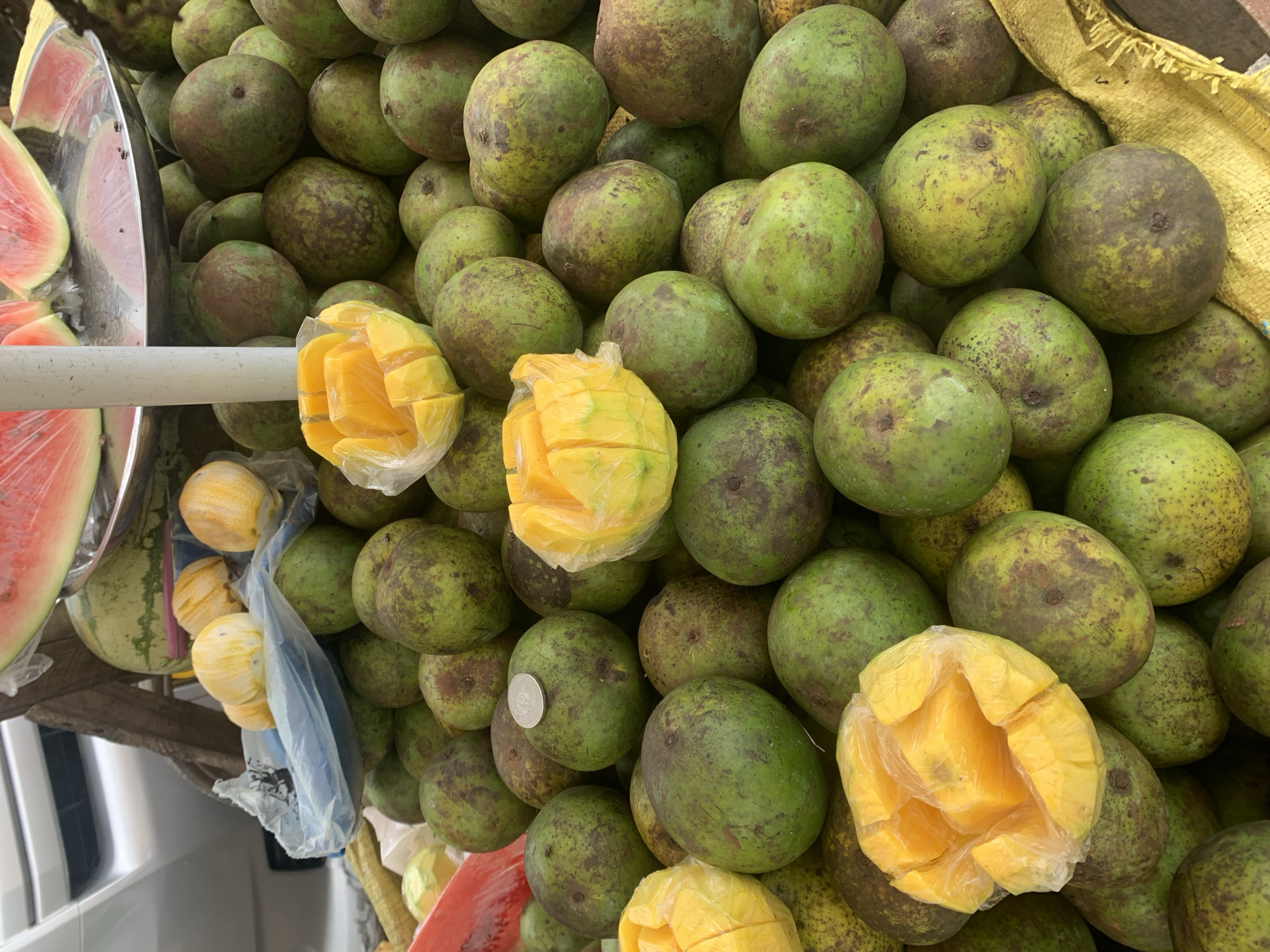 The image provides a detailed view of a fruit stall located in Dar es-Salaam, Tanzania. The primary subject is a large pile of unpeeled, green-skinned mangoes, exhibiting various shades of green with dark brown mottled markings. Interspersed within this mound are multiple bright yellow, peeled, and sectioned mangoes, each individually enclosed in transparent plastic bags. A single silver-colored coin rests on top of one of the unpeeled mangoes in the mid-ground.
In the upper portion of the frame, several slices of red-fleshed watermelon with green rinds are displayed on a reflective metallic tray. Other visible elements include a white vertical support pole, a wooden beam, and a partial view of a blue plastic bag containing pale yellow spherical fruits. A blurred silver vehicle is discernible in the upper left background, indicating an outdoor, possibly roadside, market environment. A coarse yellow woven fabric, likely part of the stall's display material, is visible in the lower right. No people are directly visible in the frame.