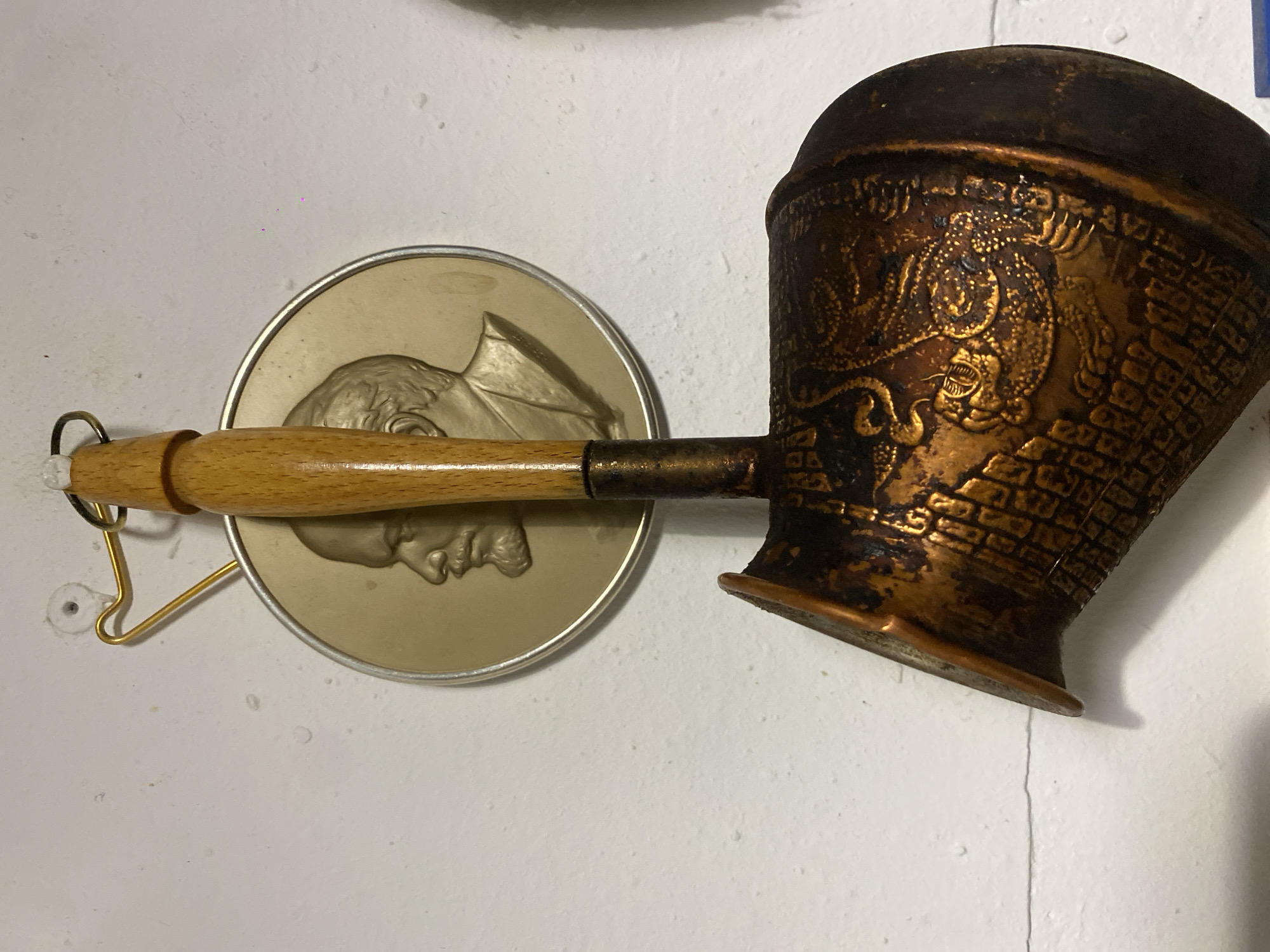 In Khimki, Russia, an indoor scene displays a metallic plaque and a traditional coffee pot (cezve) hanging against a white, textured wall. The circular plaque, situated behind the coffee pot, features a raised relief depiction of Vladimir Lenin's left profile, with a bald head and a distinct beard and mustache, wearing a suit jacket. It is composed of a brass or bronze-toned center with a silver-colored rim and is suspended by a gold-colored wire from a small, white plastic wall anchor. In front, a patinated copper or brass cezve hangs vertically. It has a light-brown wooden handle with a small metal ring at its upper end, used for suspension. The body of the cezve is adorned with an intricate embossed pattern featuring a textured, brick-like design and a stylized dragon or serpent motif in a brighter metallic tone. The lower and right portions of the cezve exhibit significant dark discoloration and patination. The white wall shows minor surface irregularities and a faint horizontal crack in the lower left quadrant.