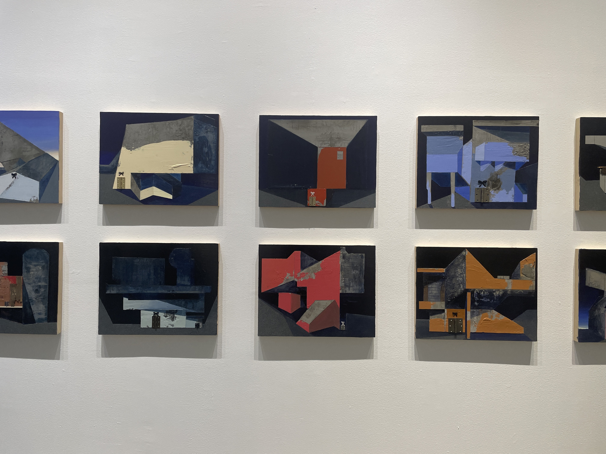 The image shows nine paintings displayed on a white wall, likely in an art gallery. The paintings are all square and abstract, with a recurring motif of geometric shapes and contrasting colors. Predominant colors include blues, grays, oranges, and reds, applied with visible brushstrokes. Each painting also features a small, almost hidden graphic of two parallel or connected rectangles, often adorned with a tiny "bow" or tie.
The location is unknown; there is no visible information in the image that confirms the Mexico City location.