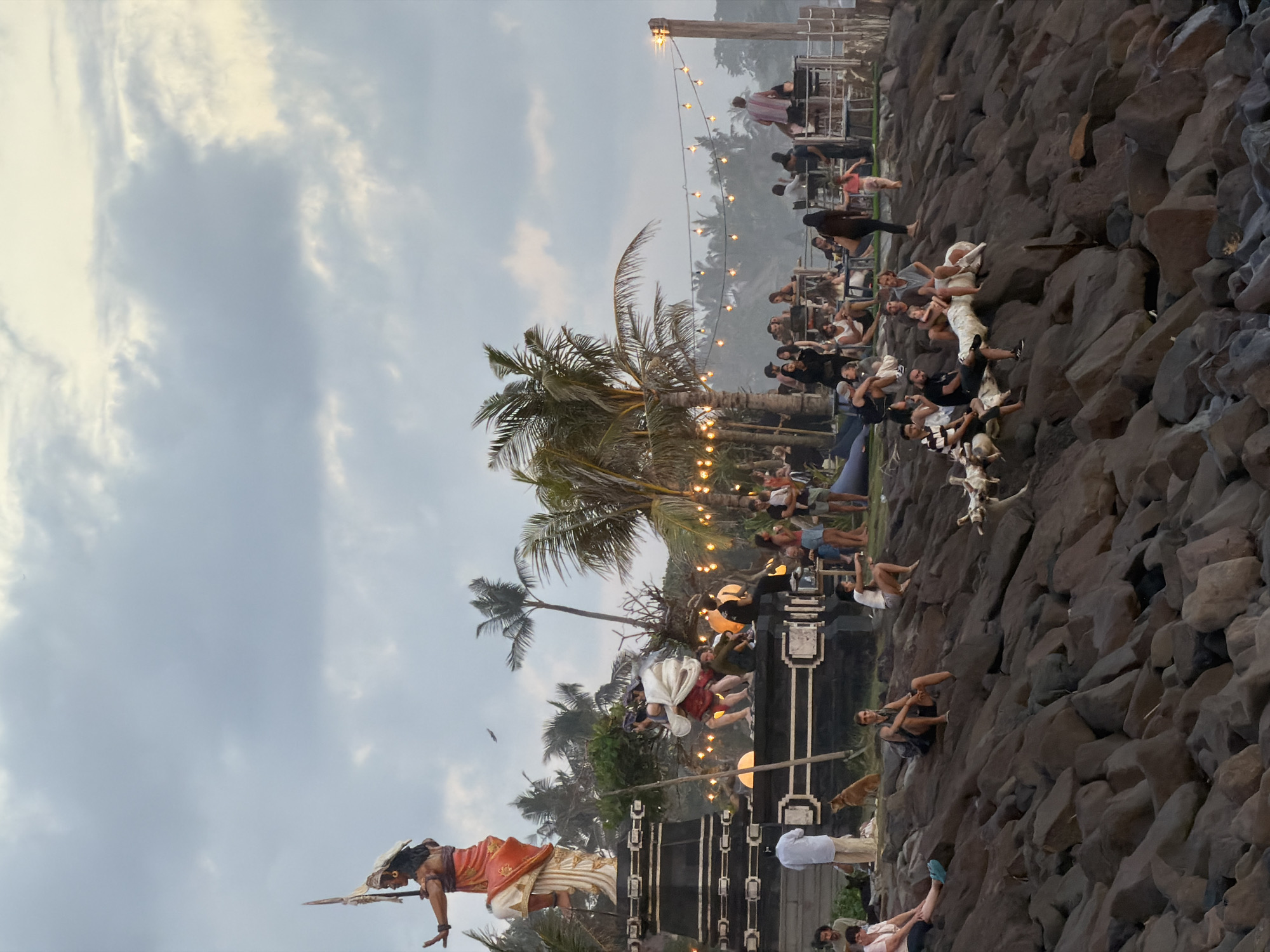 This photograph captures a lively scene at Tanah Lot, Mengwi, Indonesia. A large crowd of people is gathered along a rocky shoreline, some sitting and reclining on the rocks while others are standing further back. Palm trees with string lights intertwined in their fronds add to the atmosphere. At the foreground is a statue of a man dressed in traditional attire, standing on a platform. The sky is overcast with a mix of light and dark clouds. There are what appear to be loudspeakers on either side of the statue. String lights are draped throughout the scene.