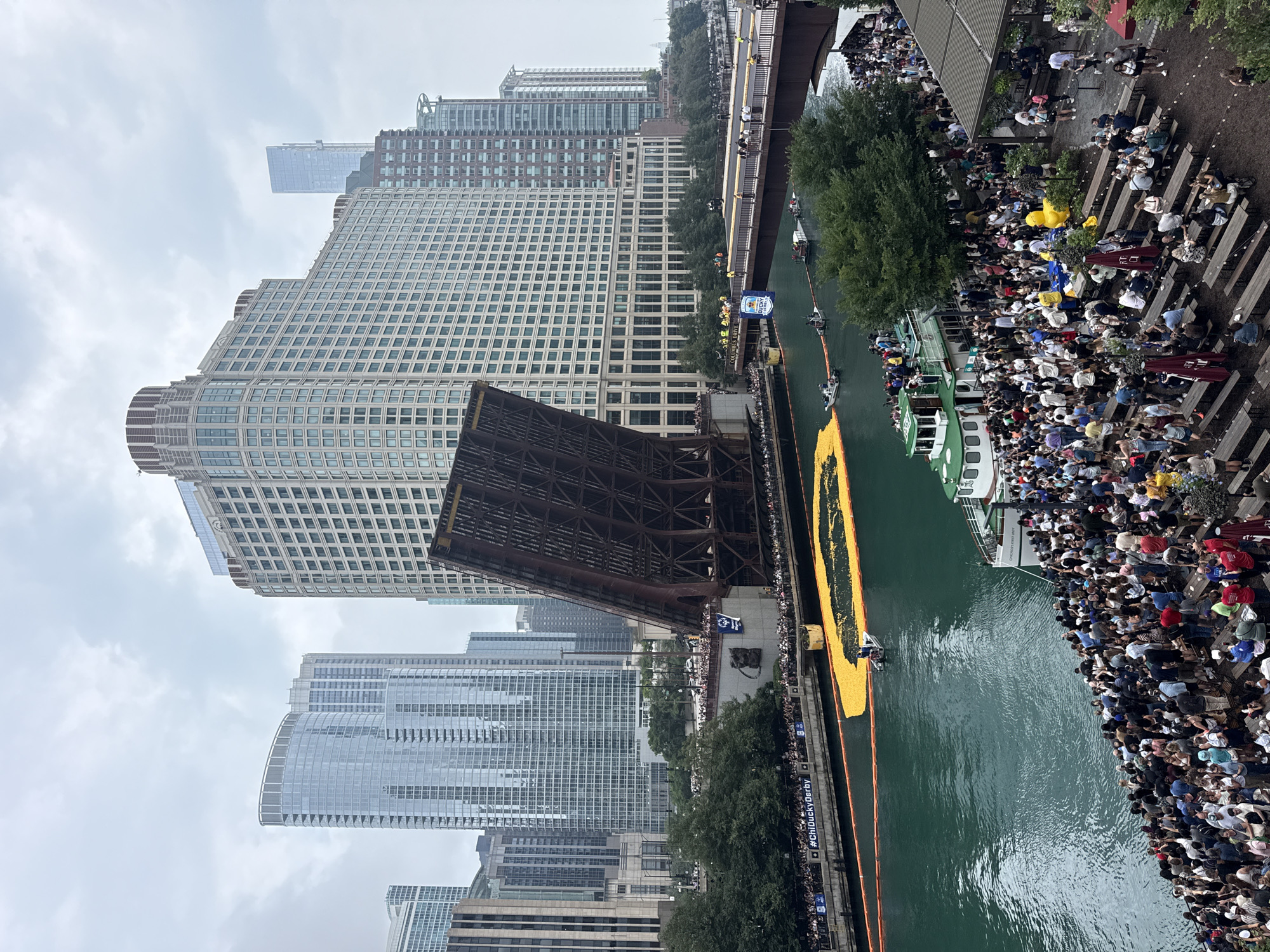 The image captures a lively scene in Chicago, Illinois, USA, likely during the Chicago Ducky Derby. The Chicago River is filled with thousands of bright yellow rubber ducks forming a floating parade, with a single small boat navigating among them. On the riverbanks and a nearby bridge, large crowds of people are gathered, observing the event. A bascule bridge, partially raised, stands prominently in the background, showcasing its intricate metal structure. Tall modern buildings, characteristic of Chicago's skyline, surround the river. A large tour boat, "Chicago's First Lady," is also visible, carrying passengers who are also watching the duck race. The overall atmosphere appears festive and energetic, with people seated at outdoor tables and standing along the river. The sky is overcast, suggesting a cloudy day.