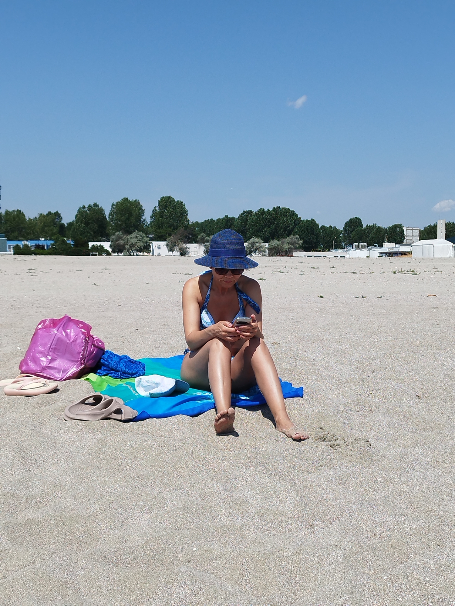 A woman is sitting on a beach towel on a sandy beach, looking at her phone. She is wearing a blue and white bikini and a blue sun hat. Beside her on the towel are a pink bag, a blue towel, and a white cap. In front of the towel, there are two pairs of flip-flops. The beach is lined with trees and buildings in the background. The sky is clear and blue, suggesting it is a sunny day. The scene appears to be a relaxing day at the beach, likely during the daytime, given the bright sunlight. The location context suggests this is Constanța, Romania. There is no visible text in the image.