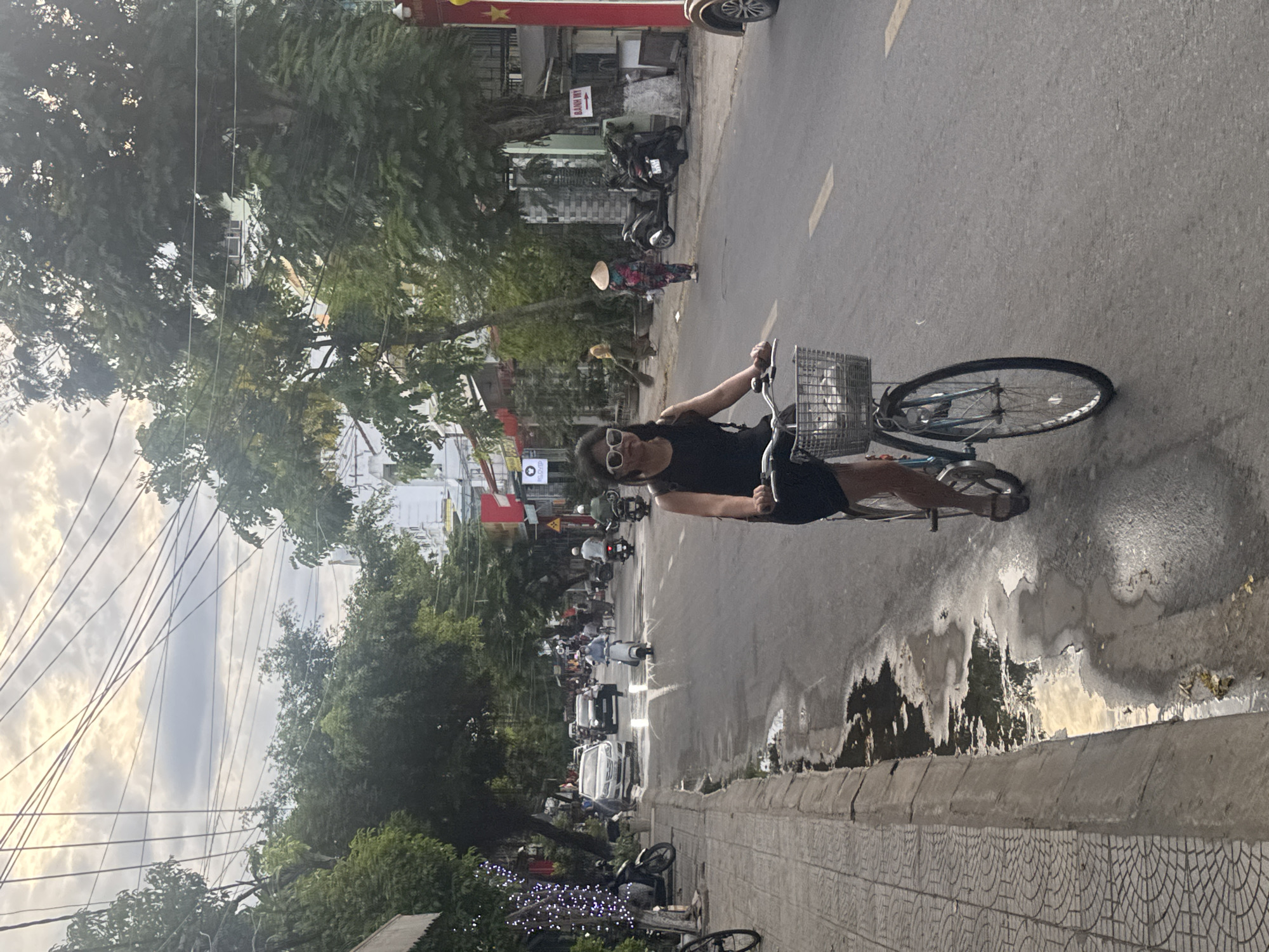 A medium shot depicts a street scene, possibly in Đà Nẵng, Vietnam, showing a woman riding a bicycle with a basket on the front, in the foreground. The woman wears sunglasses and a dark outfit. The street stretches into the background with vehicular traffic including cars and motorbikes. People are walking along the roadside. A person in a conical Asian hat is seen walking in the street, in the background. The road appears wet, with patches of standing water. The curb is visible, constructed of patterned concrete blocks. Trees and buildings line the street, and overhead power lines are strung across the sky. A red and yellow flag and sign are mounted at the roadside. The composition is somewhat vertically oriented.