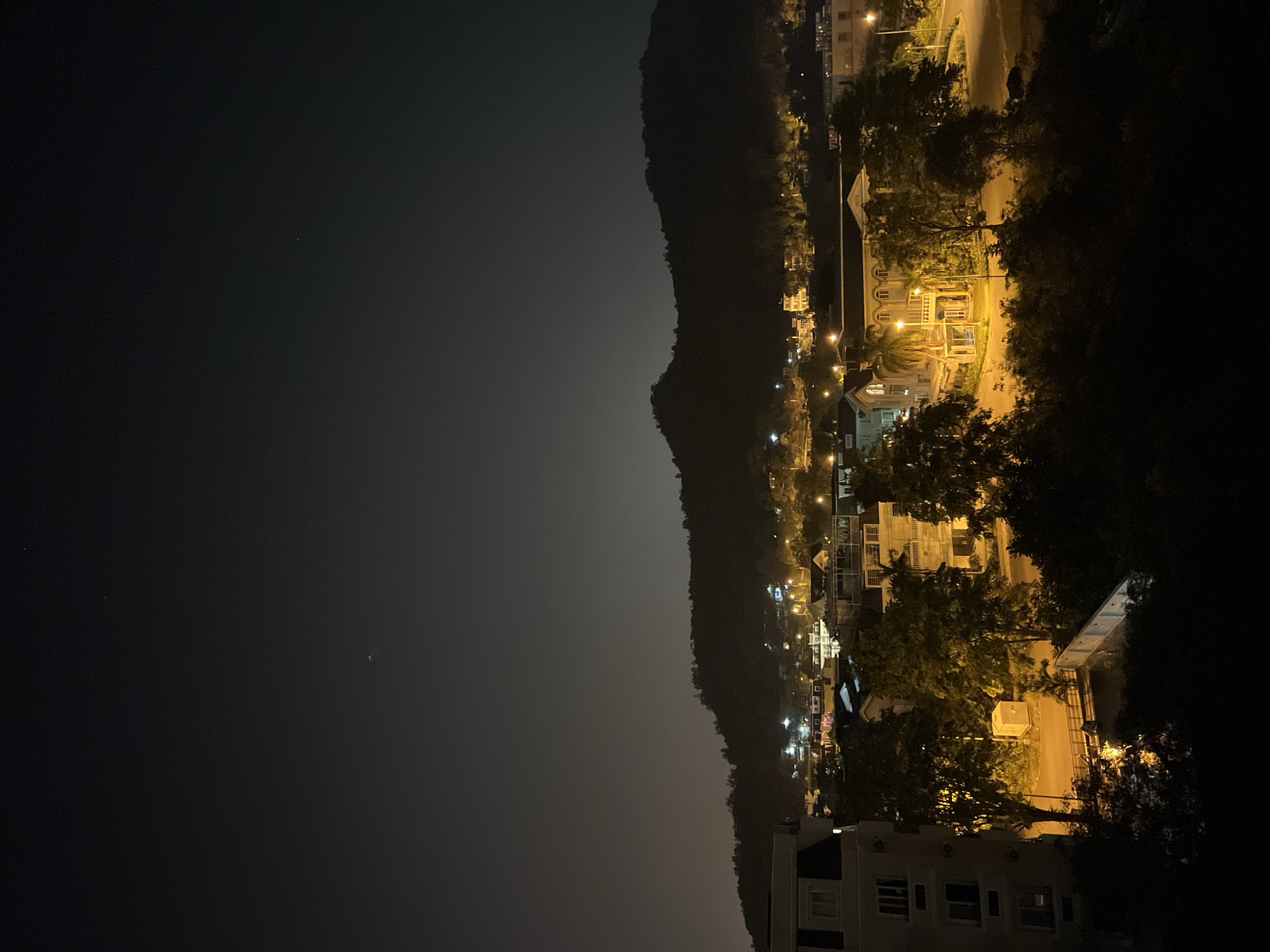 The image presents a nighttime view of Nam Nha Trang, Vietnam. A dark mountain range runs vertically on the right of the scene. To the right of the mountains is an illuminated area showing the city with buildings, street lights, and visible roads. The lights indicate residential or commercial structures along the hillside. The image is partially obscured by darkness on the left, suggesting a viewpoint either from an elevated position or overlooking a dark area. The environment has a warm tone, from the lighting. No people or actions are visible. The interaction between the landscape and the developed area is the key subject.