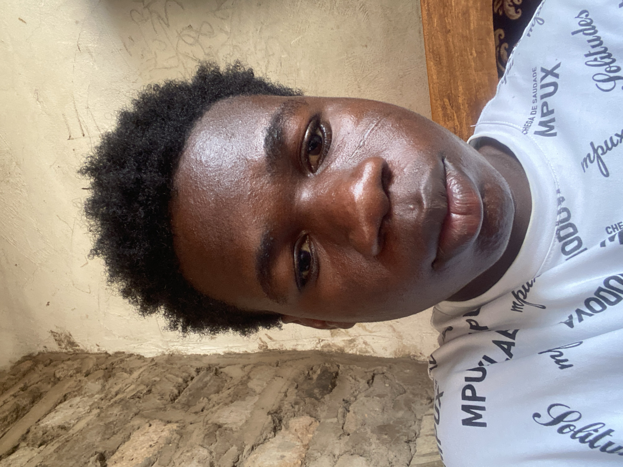 A close-up portrait, slightly tilted, features a young person with dark skin and short, curly black hair, looking directly at the camera with a neutral expression. Their face shows a noticeable sheen, possibly from sweat or skin oils.
They are wearing a white t-shirt patterned with repeating black text. Clearly visible words and phrases include "CHEGA DE SAUDADE" (Portuguese for "Enough of longing/nostalgia"), "MPUX", and "Solitudes". Other fragmented text is also present on the shirt.
The background consists of a rustic, textured wall or structure, likely made of unrendered mud or rough bricks, in shades of light brown and beige. The lower portion of the wall appears rougher and more uneven. On the right side of the image, a wooden element, possibly part of a door frame or furniture, is visible, with a glimpse of patterned dark fabric behind it. The lighting suggests it is daytime. The setting appears to be an indoor or semi-enclosed outdoor space, consistent with the provided location context of Mararaba, Nigeria.