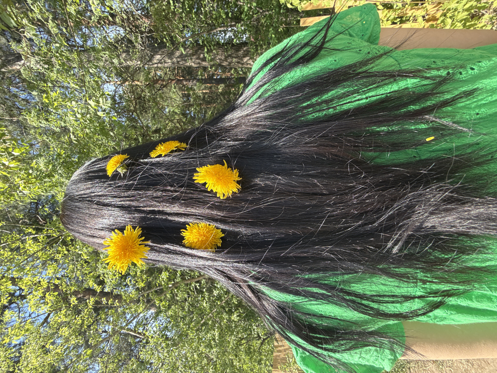 The image shows a person lying on a bright green cloth in an outdoor setting. The person has long, dark hair, and several yellow dandelions are placed in their hair. The background features trees and foliage, suggesting a park or garden. The image does not contain enough information to determine a precise location within the city of Votkinsk, Russia.