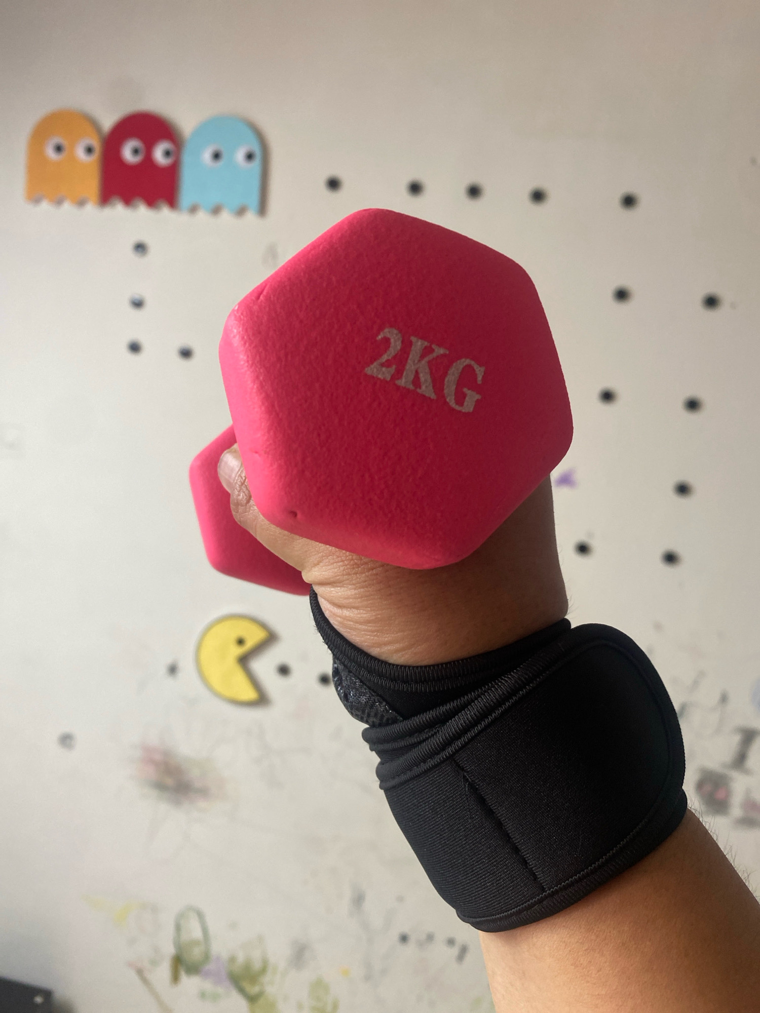 A close-up shot shows a person's arm and hand holding a pink dumbbell, which is labeled "2KG". The arm is wearing a black wrist brace. The background is a white wall with several Pac-Man themed ghost and pellet decorations. There are also some faint smudges and markings on the wall, suggesting it might be a climbing wall. The lighting is bright and direct, casting minimal shadows. There is no information about the location in Bandar Lampung, Indonesia.