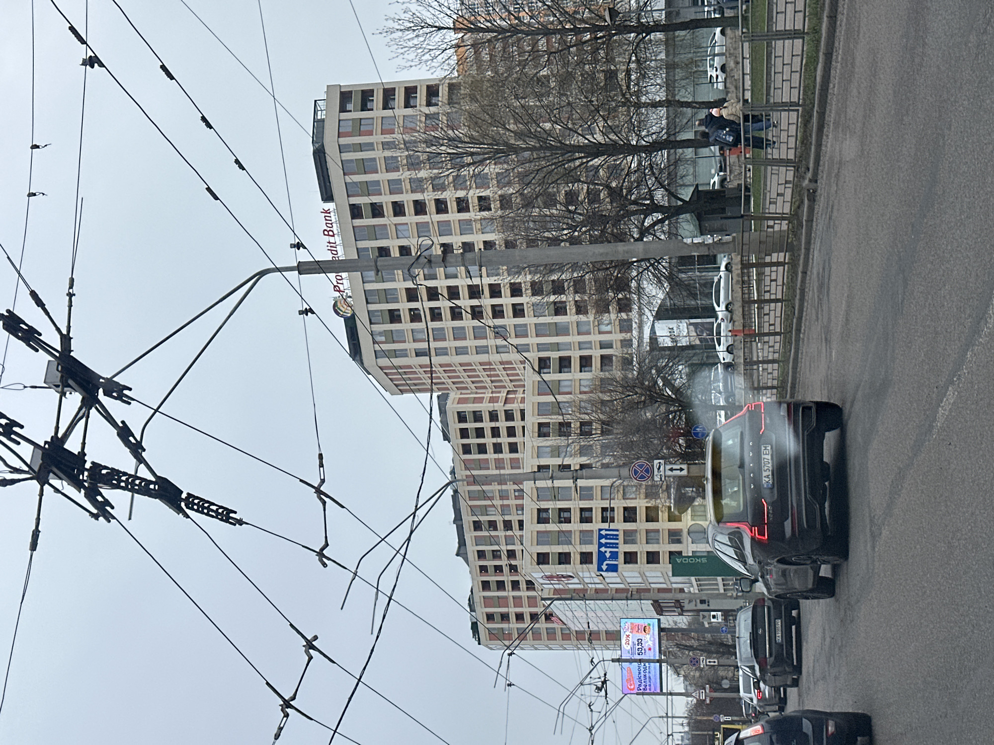 Here's a detailed description of the image:
**General Scene:** The image captures a street scene in what is likely Kyiv, Ukraine, based on the Cyrillic script on the advertisements and license plates. The perspective is from within a vehicle, looking forward along a street with moderate traffic.
**Objects and Infrastructure:**
* **Buildings:** A prominent multi-story building with a repeating grid of windows dominates the backdrop. There's signage on the building that appears to read "Procredit Bank."
* **Road:** The street is paved and shows signs of wear. Traffic is moving in both directions, but primarily in the direction of the camera.
* **Traffic:** Several vehicles are present in the foreground, including a black Volvo SUV with the license plate "KA 5707 EM." Other cars are visible ahead in the lane.
* **Overhead Cables:** A complex network of electrical or tramway cables stretches across the sky, supported by metal poles.
* **Street Furniture:** There are street signs and advertisements lining the road. A Skoda dealership sign is also visible.
* **Sidewalk:** A sidewalk is visible on the right, with a metal railing separating it from the street.
* **Trees:** Bare trees are scattered along the sidewalk.
**People:**
* A pair of figures walk along the sidewalk.
**Signage and Script:**
* Advertisements are visible, with Cyrillic text. One advertisement seems to reference "Радісного Великодн" (Likely a reference to a joyful Easter). Another sign appears to advertise a discounted price (59,99).
* The Volvo SUV in the foreground has a Ukrainian license plate ("KA 5707 EM").
* The building sign clearly states "Procredit Bank."
**Environment:**
* The sky is overcast, suggesting a grey day.
* The overall impression is a typical urban street scene.
**In summary:**
The image depicts a street in Kyiv, Ukraine, featuring a mix of commercial buildings, traffic, and street infrastructure under overcast skies.