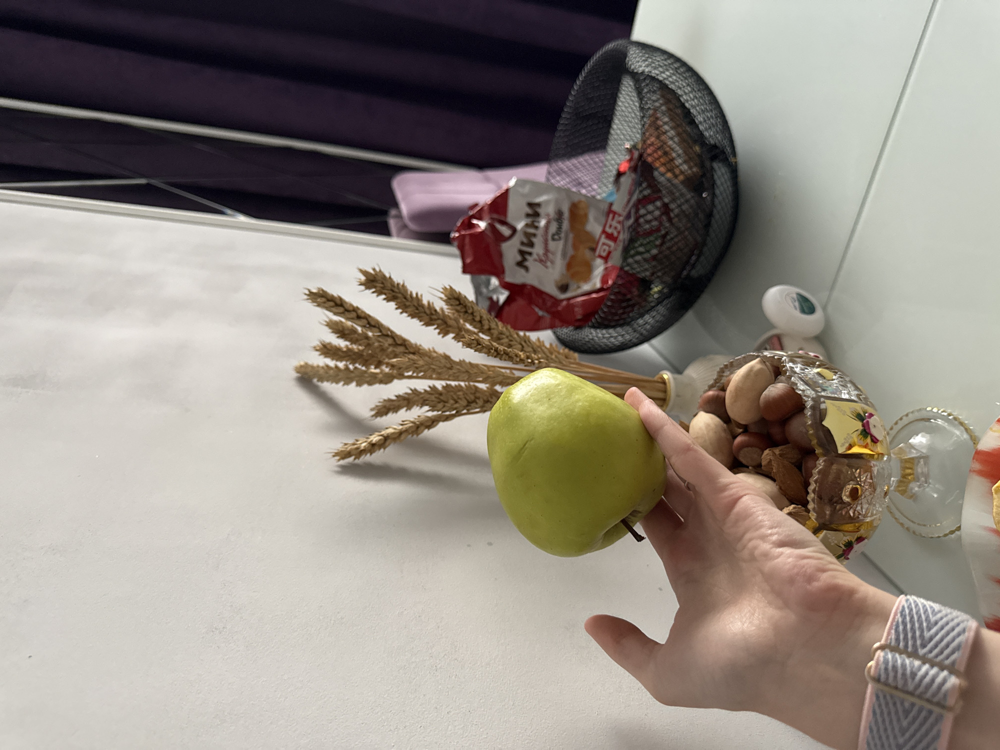 The image shows a white surface with various objects arranged on it. In the foreground, a hand with a grey and pink watch band holds a green apple. Next to the apple are stalks of wheat, a metal basket, a red and white package of cookies, a decorative bowl filled with nuts, a white object with a green circle, and a decorative item with red and white elements. A purple curtain is visible in the background.
The image does not contain information about the city of Unknown or any people.