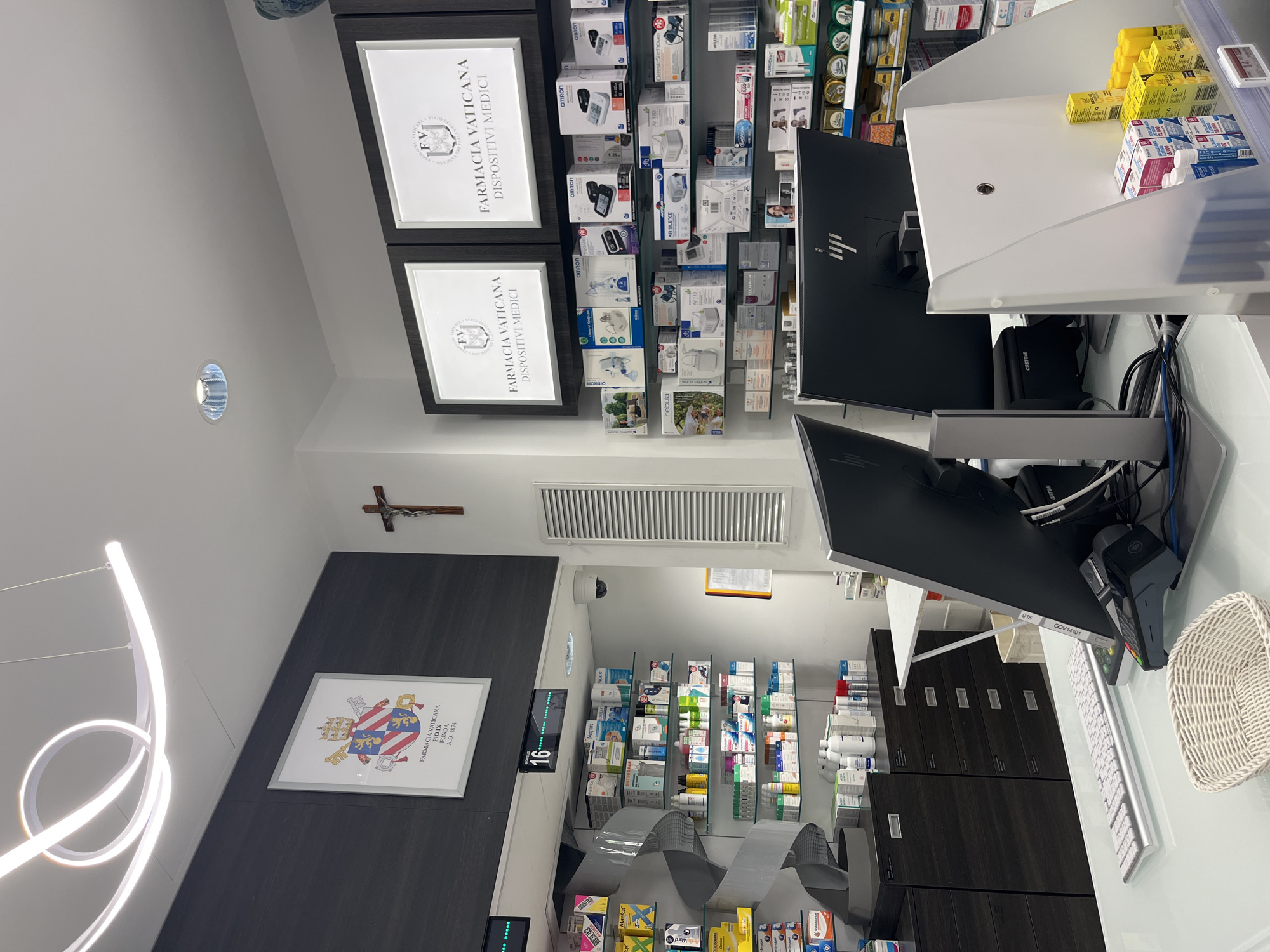 The image depicts the interior of a pharmacy, identified as Farmacia Vaticana, located in Città del Vaticano, Vatican City. The scene is well-lit with a combination of ceiling lights and illuminated signs.
In the foreground, a white counter is visible, with a white basket, a keyboard, a payment terminal, and a computer monitor with an HP logo on its stand. Another monitor is partially visible to the left. Behind the counter, shelves are stocked with various pharmaceutical products and medical devices, including boxes of medications, bottles, and packaged items. Some shelves are illuminated from behind, highlighting the products.
On the wall behind the counter, two illuminated signs display "FV" logos and text that reads "FARMACIA VATICANA DISPOSITIVI MEDICI". To the left of these signs, a framed crest with the text "FARMACIA VATICANA PIO X FONDA A.D. 1874" is displayed. A wooden crucifix hangs on the wall between the crest and the illuminated signs. A security camera is mounted on the ceiling to the right of the crest.
Further to the left, a dark wall features a spiral-shaped display or shelf unit containing more products. A digital display shows the number "16" with illuminated green dots. A ventilation grille is visible on the right wall.
The overall environment suggests a clean, organized, and professional pharmacy setting within the Vatican City. There are no people clearly visible in the image.
