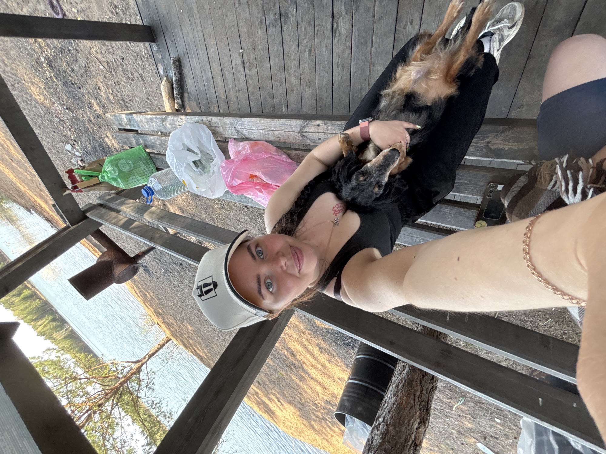 A rotated upward selfie photograph captures a woman and a dog on a wooden structure, possibly a deck or gazebo. The woman, wearing a black tank top and a white baseball cap, is holding the dog in her arms. The dog is small to medium-sized, with black and brown fur. The woman is also wearing a necklace, a bracelet, and sneakers.
Beside them, on the wooden surface, are various items, including plastic bags in white, green, and pink, and what appears to be a clear plastic bottle. The background shows a landscape with water and trees. The photograph seems to have been taken in Selychka, Russia, which appears in the geolocation data of the photograph.