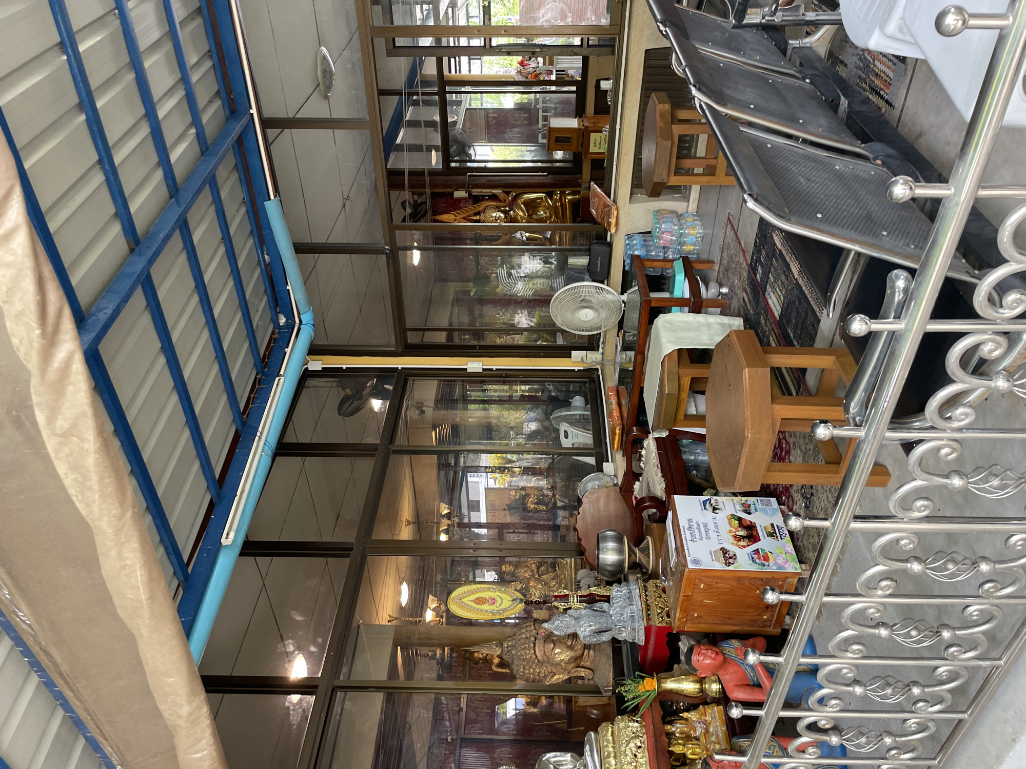 The image shows an indoor area of a temple or shrine in Tambon Na Chom Thian, Thailand. The space is covered by a blue-framed metal roof with a corrugated metal ceiling, and is enclosed by large glass windows. The interior is filled with various religious artifacts and furnishings. In the foreground, a metallic railing with decorative scrollwork separates the viewer's area from the main space. Beyond the railing, there are several small wooden tables and octagonal wooden stools. A fan is visible to the left, and to the right, a row of black plastic chairs with metal frames suggests a waiting or seating area.
On display are multiple Buddha statues of varying sizes and materials, including gold-colored ones and stone-carved figures. One large Buddha head statue is prominent. There are also decorative fans, vases with flowers, and other religious objects. A wooden donation box with informational flyers is placed on a small wooden cabinet. The floor is covered with patterned rugs. Through the glass windows, more religious imagery and the interior of the temple are visible, including golden Buddha statues. The overall impression is of a well-maintained and decorated religious site.