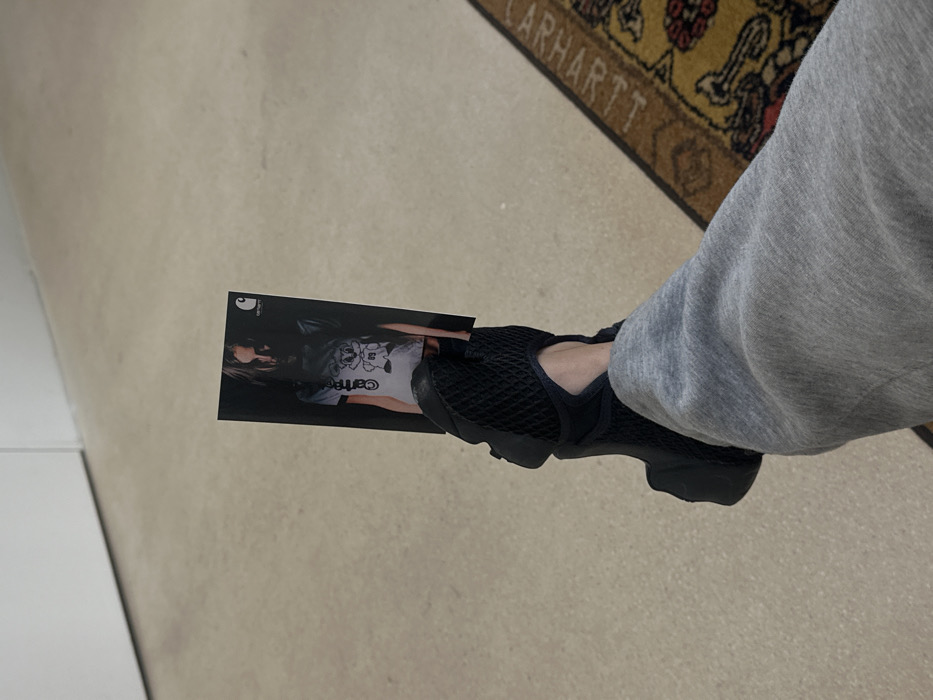 The image presents a close-up, first-person perspective of a person's lower legs and feet. The individual is wearing grey sweatpants and black, mesh-textured athletic shoes. A rectangular promotional card is held vertically by the left foot, wedged between the shoe and the foot itself. This card features a dark-haired model wearing a light-colored t-shirt with a cartoon character and the number "99" on it, along with visible "Carhartt" branding on the shirt and the "Carhartt WIP" logo in the upper right corner of the card. The surrounding environment consists of a light beige floor, a plain white wall in the upper left, and a segment of a brown, yellow, and red patterned rug with "CARHARTT" text along its border, visible on the right side of the frame. Based on the prominent and repeated Carhartt branding, the scene is located within a Carhartt Work In Progress (WIP) retail store in Paris, France.