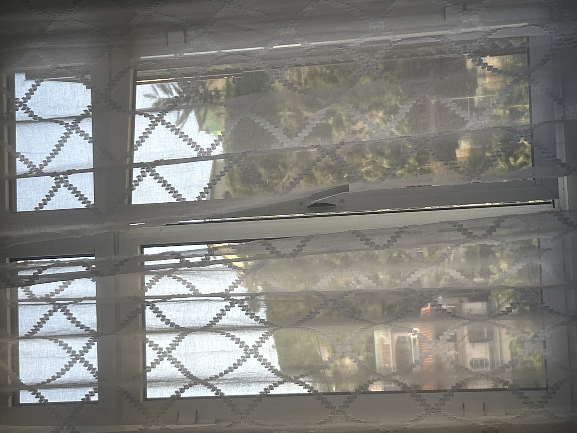 The image shows a view through a window covered with a white patterned curtain. The window is divided into multiple panes. Outside, there are trees and foliage, and a glimpse of buildings. There is no discernible information about the location of Câmara de Lobos, Portugal, or any people, subjects, actions, or interactions.