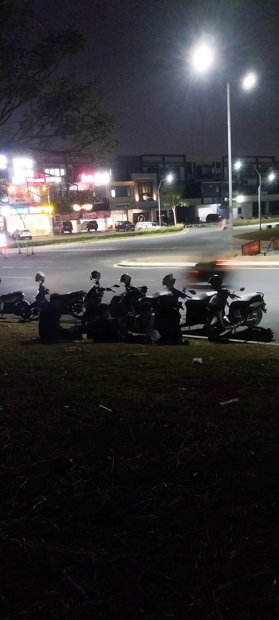 The image depicts a nighttime scene on a street in Medan, Indonesia, characterized by artificial lighting and the presence of parked motorcycles and several individuals. The foreground is dominated by a grassy area with several motorcycles parked in a row. Some individuals appear to be sitting or standing near the motorcycles. Beyond the grassy area, a paved road curves into the distance, with vehicular traffic indicated by motion blur. On the opposite side of the road, a row of modern buildings with illuminated storefronts and windows is visible. Streetlights illuminate the scene, with one particularly bright light source in the upper right quadrant of the image. The overall atmosphere suggests a commercial or residential area at night.