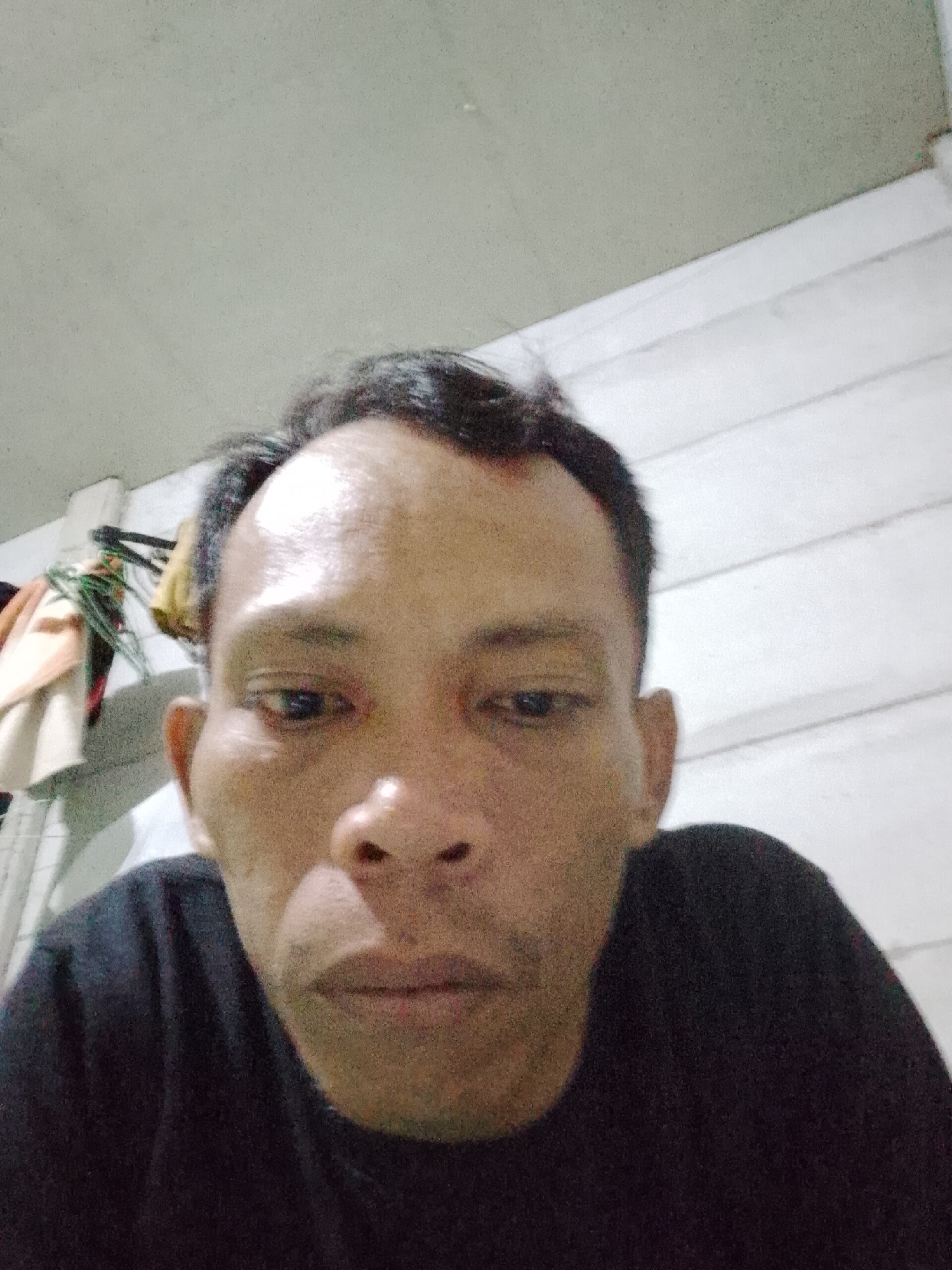 A medium close-up shot shows a man wearing a black t-shirt, looking slightly down at the camera. The background appears to be an interior space with off-white walls and wooden panels. Hangers and clothing are visible on the left side of the frame. The location is Bukit Ketok, Indonesia.