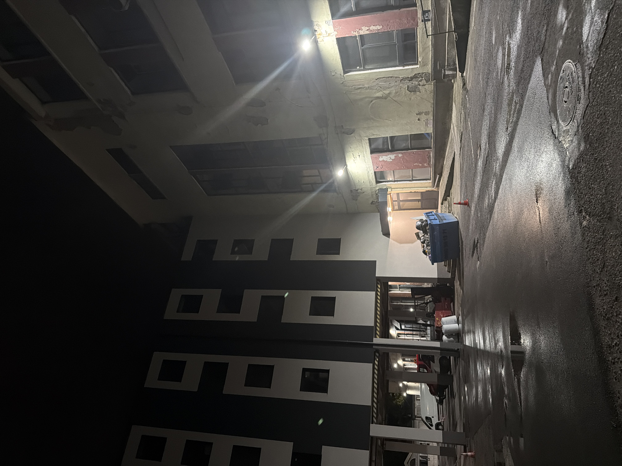 The image depicts an exterior view of a residential building complex at night, possibly in Plovdiv, Bulgaria. The scene is illuminated by artificial lights, casting reflections on a wet asphalt ground surface marked by puddles. In the foreground, a manhole cover is visible. To the left, a modern building with a dark and white facade featuring geometric window patterns stands. Adjacent to it, a covered walkway with white pillars and yellow and black striped beams is present, sheltering parked vehicles including a white van and a red truck, as well as two large white sacks. Further to the right, an older building with a beige facade exhibits signs of wear and tear, with peeling paint around several windows, some of which have red shutters. A blue dumpster filled with debris is situated in front of this older building, near an orange traffic cone and a ramp. Two streetlights are visible on the facade of the older building, casting bright beams. The overall atmosphere is dimly lit and appears to be a service or common area of the complex. No people are present in the frame.