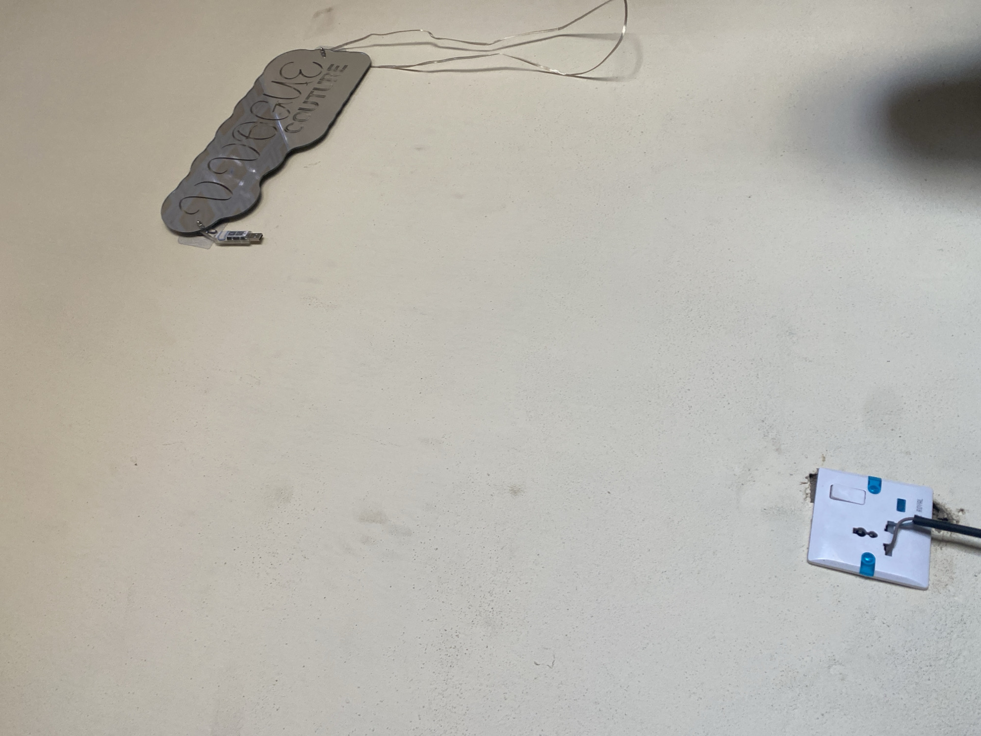 This is a forensic, matter-of-fact description of an image, focusing on objective details. The image depicts a section of a lightly textured, off-white wall. In the lower left quadrant, a white electrical socket is mounted. The socket has three distinct openings and two small blue indicators. A black cable is connected to the socket and descends downwards. Above and to the right of the socket, an illuminated sign made of a silver, reflective material is attached to the wall. The sign is elongated and wavy in shape, with cut-out lettering that reads "VOGUE COUTURE." A thin, metallic wire is attached to the sign and trails downwards. A small, rectangular USB drive is also visible, plugged into a port near the top of the sign. The lighting in the scene creates a shadow on the lower right portion of the wall. The overall environment appears to be indoors, possibly in a commercial or residential setting. There are no people or animals present in the image. The location is identified as Jauro Yinu, Nigeria.