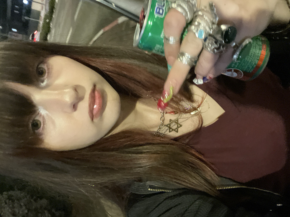 A woman with long brown hair and bangs poses while holding a green can. She wears a burgundy top and a black jacket. Several silver rings adorn her fingers, and she has long, colorful fingernails. A silver necklace with a Star of David pendant hangs around her neck. The background shows a dimly lit street scene.
