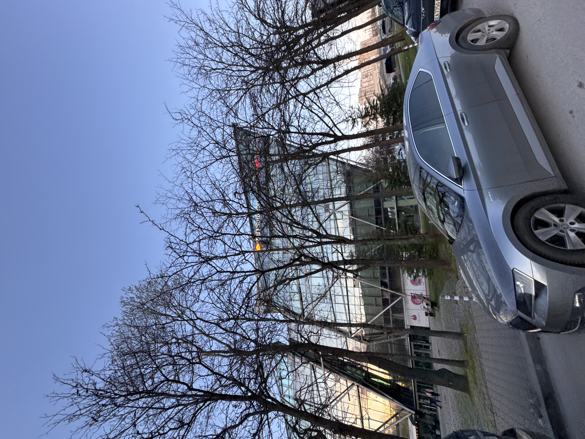 The image shows an outdoor scene in Sofia, Bulgaria. The foreground features a gray car parked on a paved surface, presumably a street or parking area. Behind the car are leafless trees that partially obscure a modern glass building. The building appears to be a commercial or office structure with a visible sign, likely displaying the name of a company or organization. The sky is a clear blue. The overall impression is of a modern urban environment.