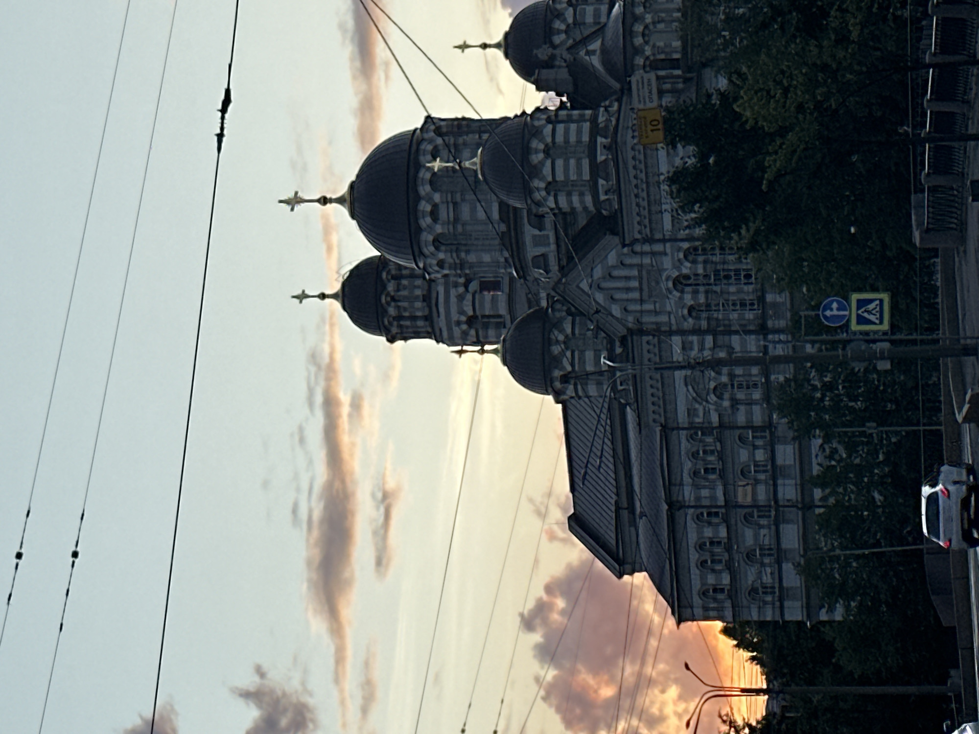 The image captures a view of the Saint Nicholas Naval Cathedral (Nikolskiy morskoy sobor) in Saint Petersburg, Russia, during a sunset. The cathedral, with its striking blue and grey facade and multiple domes topped with crosses, dominates the frame. Overhead, power lines crisscross the sky, with some reflecting the soft hues of the sunset. In the foreground, a road is visible with traffic, including a white car driving away from the camera and street lights illuminating the scene. Trees line the embankment, partially obscuring the lower part of the cathedral. A traffic sign with a number 10 and a yellow sign are also visible on the cathedral's facade. The sky displays a gradient of soft oranges, pinks, and blues, indicative of the twilight hour.