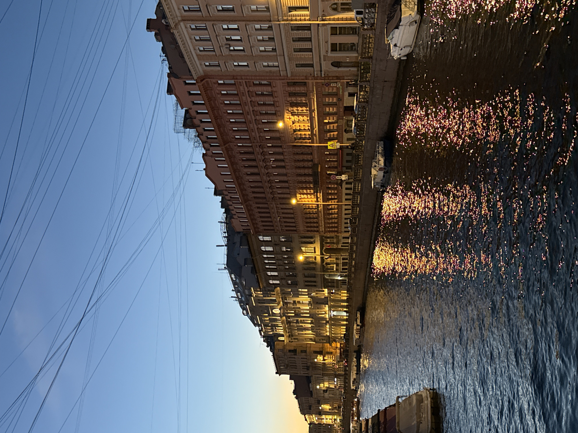 This image captures a canal in Saint Petersburg, Russia, at dusk. The scene is dominated by a row of ornate, old buildings lining the canal, their facades illuminated by warm streetlights. The buildings exhibit classical architectural styles with numerous windows and intricate detailing. Boats are docked along the canal's edge, and a bridge can be seen in the distance.
The canal's dark water reflects the lights from the buildings and the sky, creating shimmering patterns. The sky transitions from a clear blue in the upper left corner to a soft orange-yellow near the horizon, indicating the setting sun. A network of thin, dark wires crisscrosses the sky, a common sight in many urban environments. The overall mood is serene and picturesque, evoking a sense of historical charm and urban tranquility. No people are distinctly visible. The time of day is clearly twilight, and the weather appears to be clear.