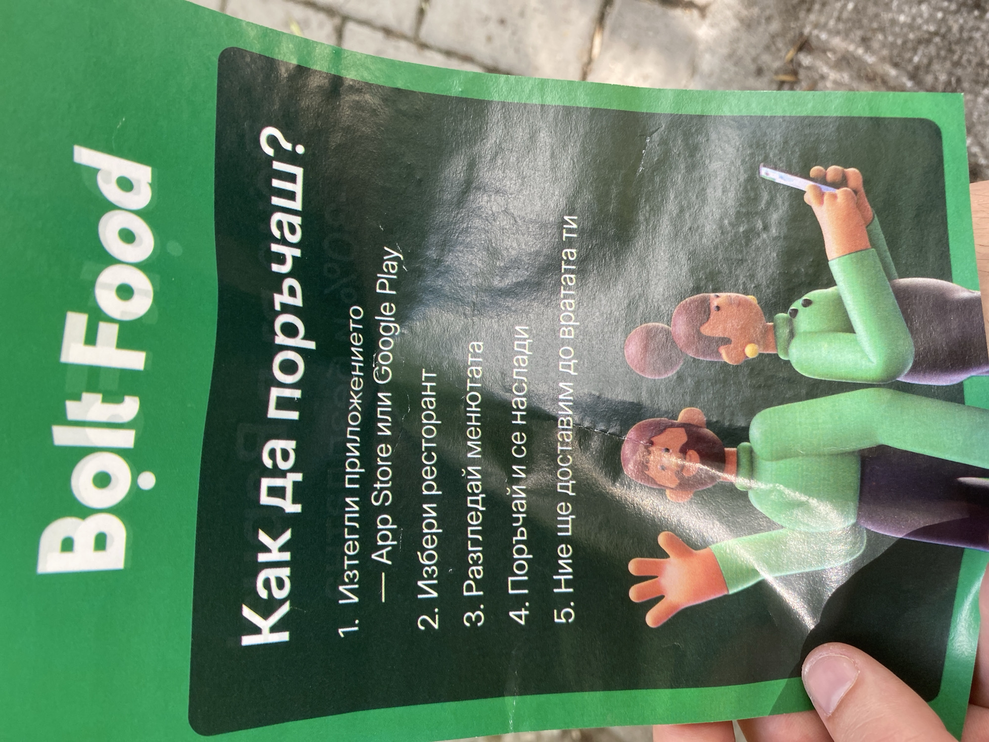 The image shows a flyer with the Bolt Food logo prominently displayed at the top, followed by instructions in Bulgarian on how to order food. The text translates to: "How to order?". The numbered steps are: 1. Download the application - App Store or Google Play, 2. Choose a restaurant, 3. Browse the menu, 4. Order and enjoy, 5. We will deliver to your door. The flyer also features cartoon-like illustrations of a man and a woman. The man is waving with his left hand, and the woman is holding a smartphone. The background is a mottled green with a faint watermark. The flyer is held by a person's hand on the left side of the image. The background outside the flyer appears to be a paved surface, possibly a sidewalk or street, with some debris visible. The image was taken in the city of Varna, Bulgaria.