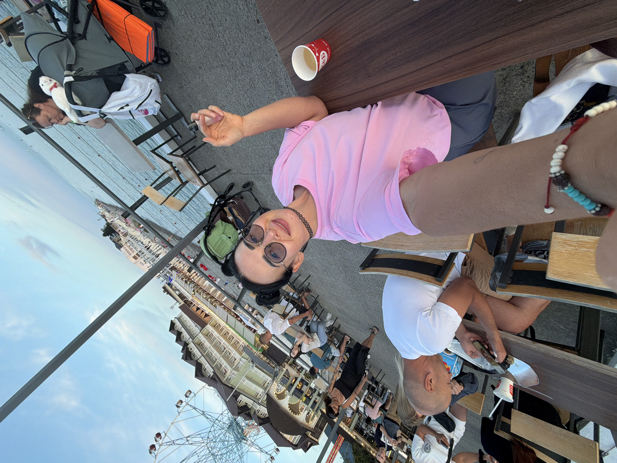 The image depicts a group of people at an outdoor cafe or restaurant, likely near a body of water, with a ferris wheel visible in the background. The scene appears to be a leisure setting, possibly a seaside resort or boardwalk. A woman in a pink shirt is prominently featured in the foreground, taking a selfie. Other individuals are seated at tables, some eating or drinking. The environment includes tables, chairs, umbrellas, and a railing. The sky is visible with some clouds.