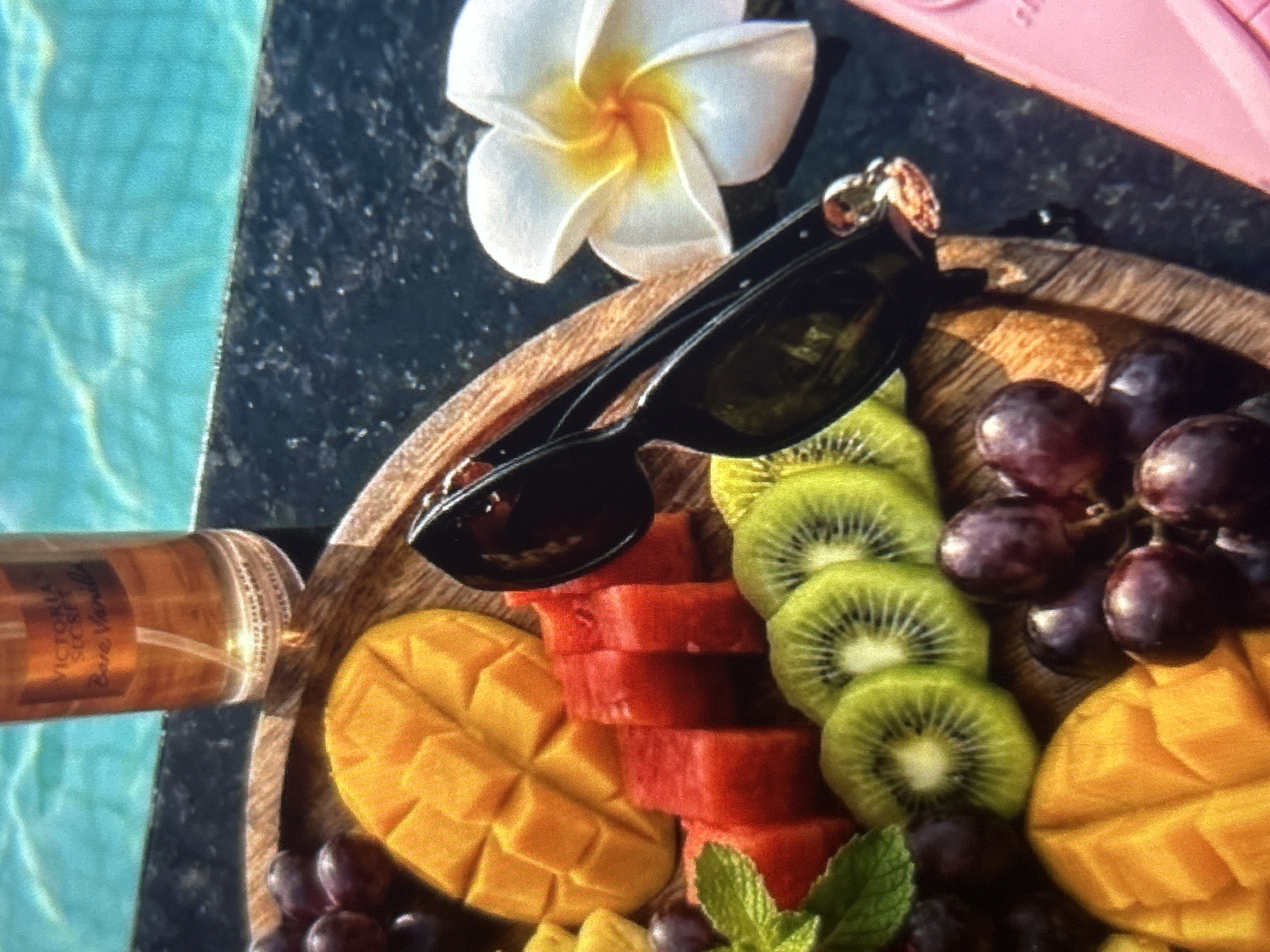 This is a close-up, slightly overhead shot of a fruit platter and other items placed on the edge of a swimming pool. The fruit platter, made of wood, contains sliced mango, watermelon, kiwi, and grapes. A pair of black sunglasses rests on the platter, angled across the fruit. To the right of the platter, a white flower with a yellow center, possibly a plumeria, is visible. In the background, the turquoise water of the swimming pool is slightly rippled. A clear glass bottle with an orange label, identifiable as a Victoria's Secret "Bare Vanilla" body mist, stands near the edge of the pool. The scene appears to be outdoors, likely in a warm climate, given the pool setting. There are no people visible in the image. The location is not specified as Knezha, Bulgaria, or any other city.