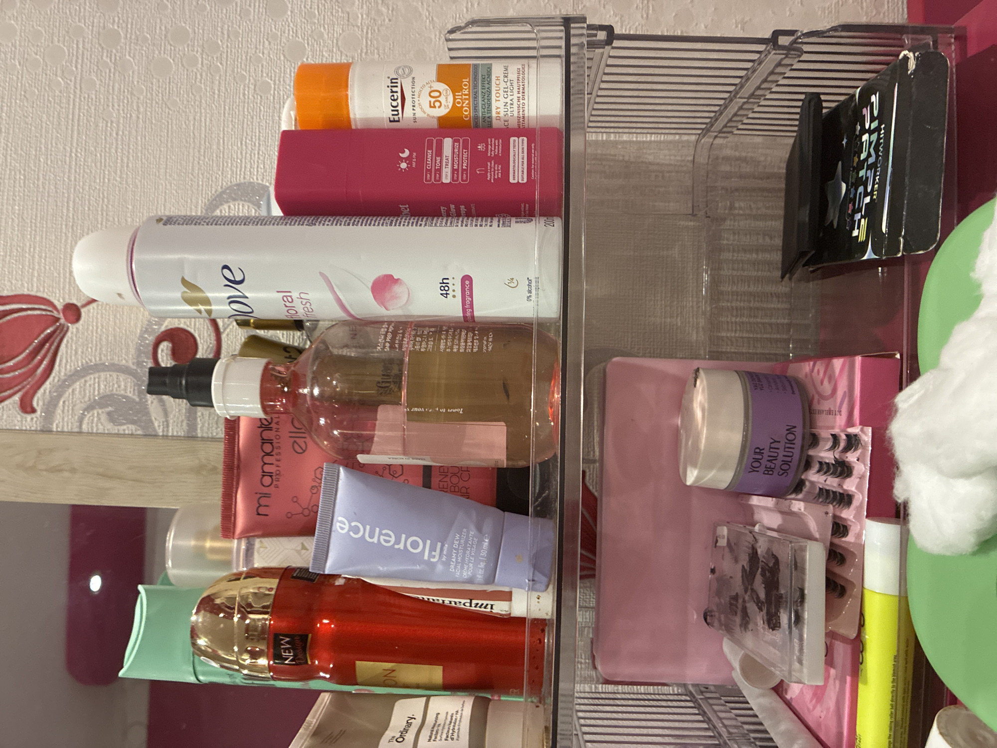 The image captures a collection of personal care and beauty products neatly organized within clear acrylic containers, likely situated on a vanity or bathroom counter. The background features a white textured wallpaper with a repeating circular pattern, accompanied by a red and pink floral motif.
In the upper-most section of the organizer, an orange and white Eucerin bottle for 'SUN PROTECTION OIL CONTROL 50+ DRY TOUCH SUN GEL-CREME ULTRA LIGHT' is visible. Adjacent to it is a pink tube with instructions for a skincare routine: 'CLEANSE, TONE, TREAT, MOISTURIZE, PROTECT' and 'Glow drops'. Below these items, a white Dove aerosol deodorant spray, labeled 'floral fresh 48h 0% alcohol', is prominently displayed.
The middle section holds a clear bottle with a pink label indicating 'mi amante PROFESSIONAL elle'. Beside it, a lavender-colored tube from 'florence by mills' is identified as 'DREAMY DEW FACIAL MOISTURIZER CREME HYDRATANTE POUR LE VISAGE 30 ml'. A striking red and gold bottle, marked 'NEW Design' and 'Imparat', and a beige tube of 'The Ordinary Natural Moisturizing Factors +HA' are also part of this organized display.
On the right side, within a separate clear compartment, a purple jar labeled 'YOUR BEAUTY SOLUTION YUZ BAAR Sebum' is visible. Below it, several trays of false eyelashes are meticulously arranged. A black compact or palette shows text including 'SIMPLE HITWORKER'. A yellow stick, possibly a glue stick, and fluffy white cotton balls complete this section. The products are reflected on the underlying surface, suggesting a mirror or a glossy countertop. The overall arrangement indicates a well-maintained and comprehensive beauty and skincare regimen.