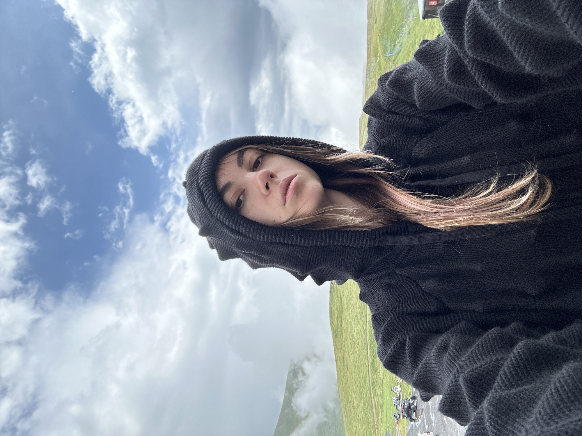 A young woman with light brown hair, some of which is dyed pink, is taking a selfie outdoors. She is wearing a black hooded sweater with a textured pattern on the hood and sleeves. Her face is turned towards the camera, and she is looking directly at it with a neutral expression. Her hair is visible around her face and falls over her shoulders.
The background of the image shows a cloudy sky with patches of blue. Below the sky is a landscape of rolling green hills, partially obscured by mist or fog in some areas. In the distance, there appear to be several ATVs parked on a wet, possibly paved area. A fence is also visible in the background. The overall impression is of a cool, possibly damp, outdoor setting in a mountainous region. The location is identified as Gudauri, Georgia.