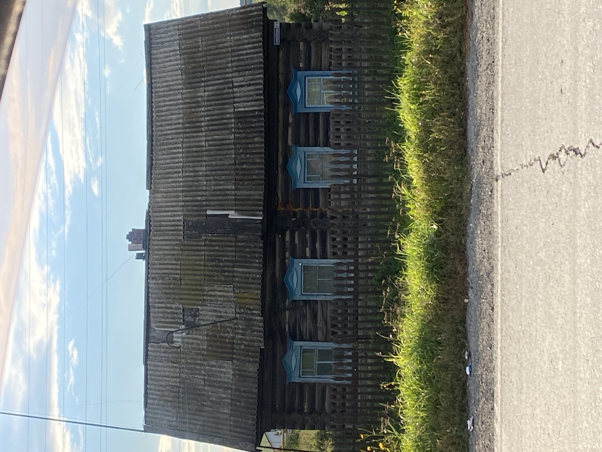 This is a front-facing, eye-level shot of a rustic, aged wooden house with a corrugated metal roof, situated in the village of Медведское, Russia. The house has four windows with decorative blue trim, and a dark wooden fence stands in front of it. Beyond the fence, a strip of overgrown green grass and some small yellow flowers are visible. The scene is framed by a road in the foreground and a cloudy blue sky with power lines overhead. The overall impression is one of rural simplicity and weathered charm.