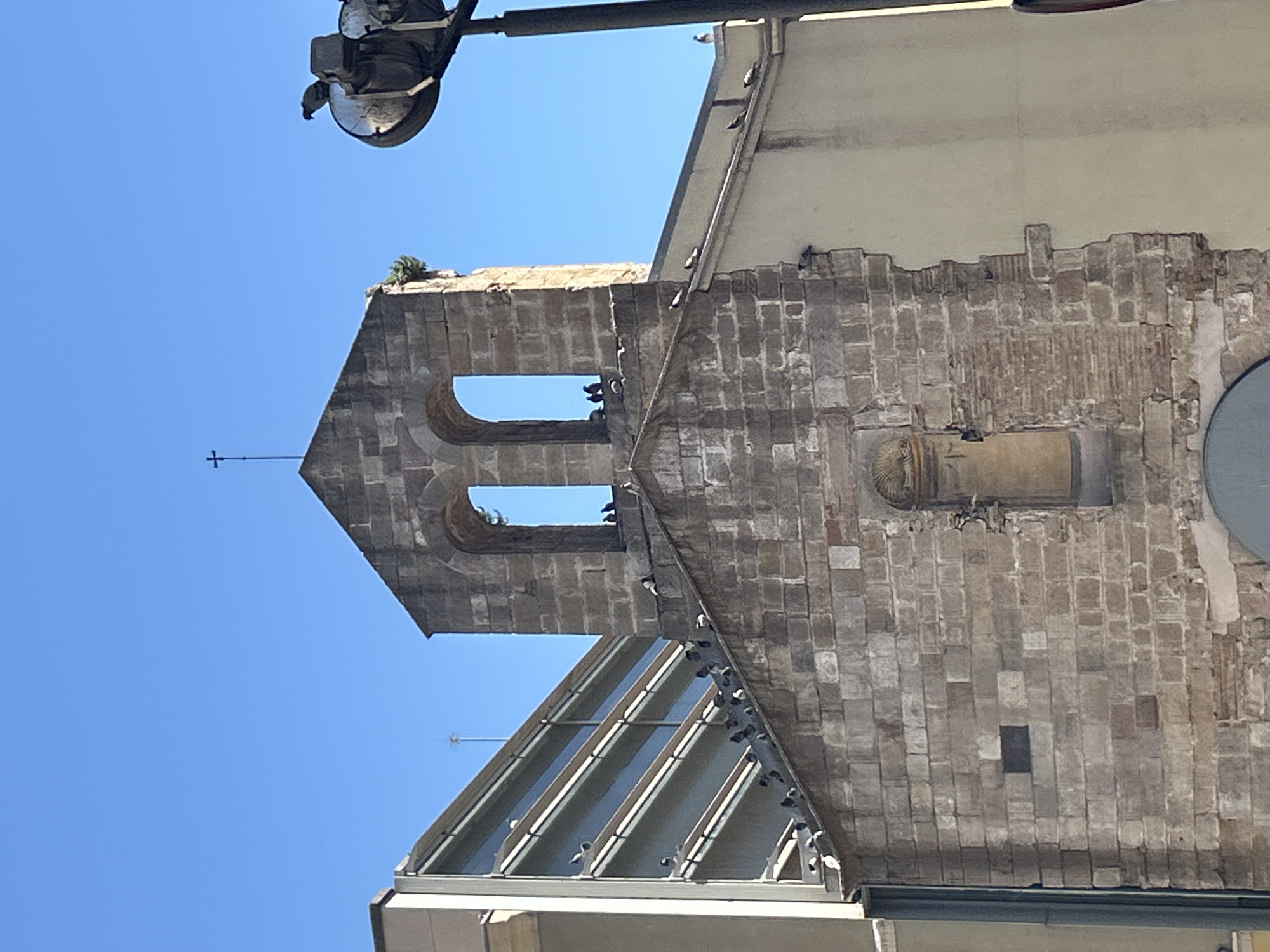 The image presents a striking architectural contrast against a clear blue sky, likely in an urban setting like Barcelona, Spain. The primary subject is an ancient stone bell tower, likely part of a church or historic building, constructed from weathered, light-colored stone blocks. This bell tower features two prominent arched openings, one positioned above the other, and is topped with a slender cross. Small bits of green vegetation grow on its upper edges, hinting at its age. A decorative niche with what appears to be a small statue or carving is visible on the right side of the tower's facade.
Adjacent to this historic structure, a section of a modern building is visible, featuring a distinctive sloping roof composed of reflective glass panels and dark metal support beams. Numerous pigeons are scattered across both buildings; they perch on the ledges and beams of the modern roof, as well as on the old stone tower and within its arched openings.
In the upper left portion of the frame, a dark, circular street light fixture extends into the clear, vibrant blue sky. The image is taken looking upwards, emphasizing the height of the structures. The bright, clear sky indicates it is daytime with strong sunlight. There is no visible text. The scene captures a peaceful moment of urban life where old and new architecture coexist, populated by resting birds.