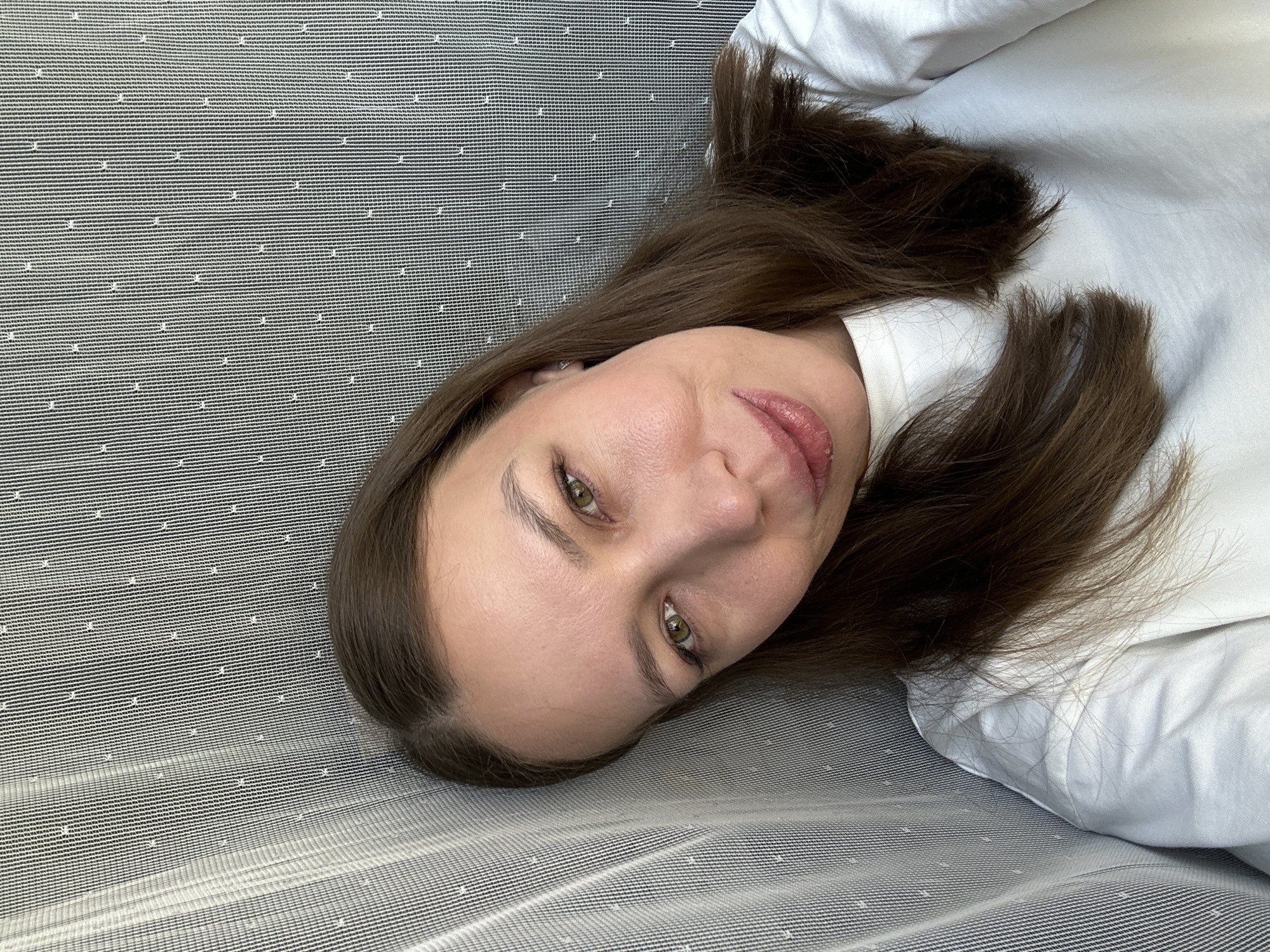 A woman with long brown hair, fair skin, and green eyes is lying on a gray and white striped surface. She is wearing a white shirt. No information indicates this image was taken in Tomilino, Russia.