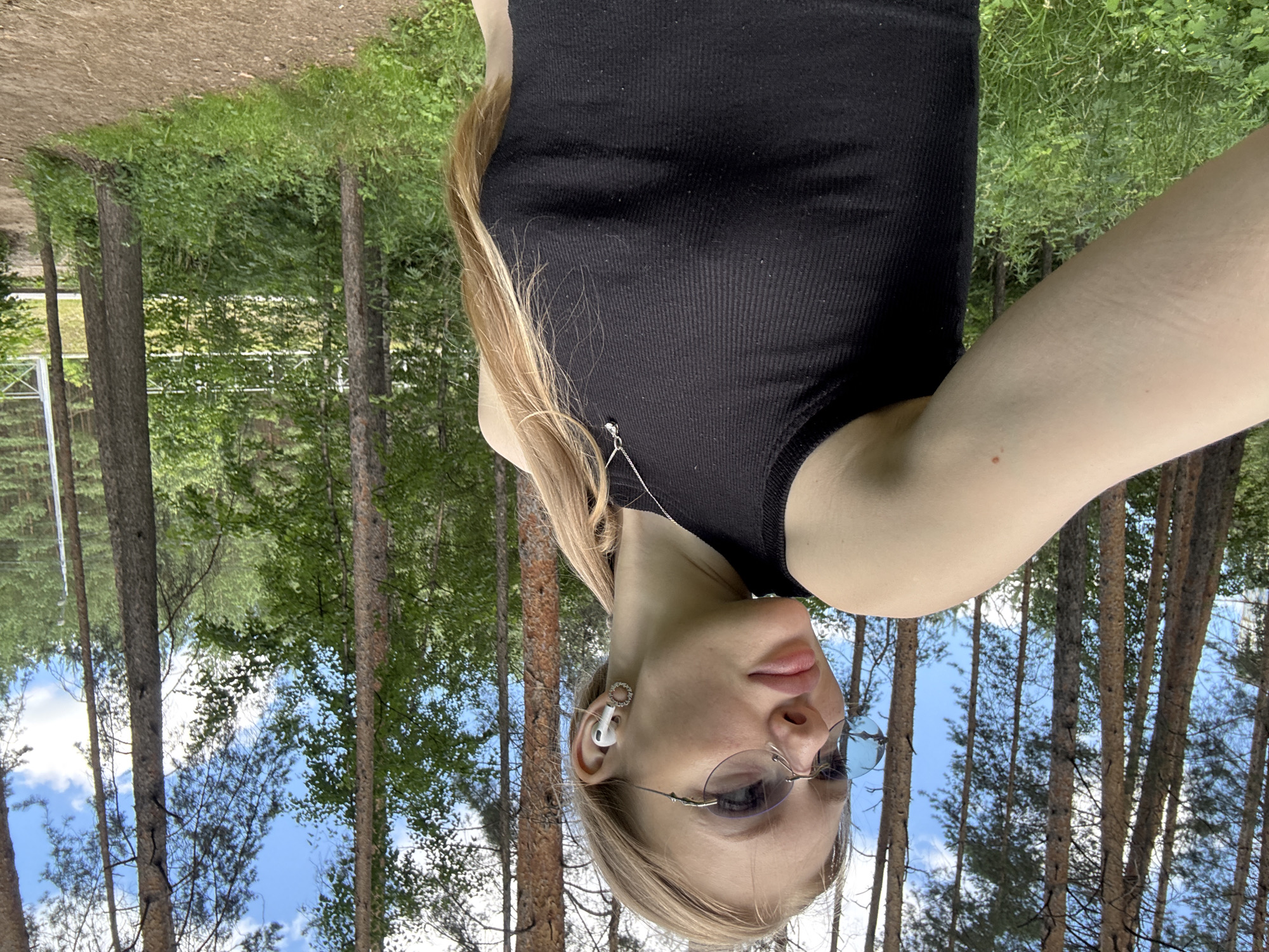 In a natural setting amidst tall pine trees and lush green undergrowth, a young woman with blonde hair pulled back in a ponytail is captured in a selfie. She is wearing a black ribbed tank top, rimless oval sunglasses, and white wireless earbuds. A delicate silver necklace with a small pendant rests on her chest. Her left arm is extended towards the camera, and her right side is visible, with the background showing a dirt path and more trees, suggesting an outdoor recreational area. The sky above is partly cloudy with patches of blue. The location is identified as the city of Perm, Russia.