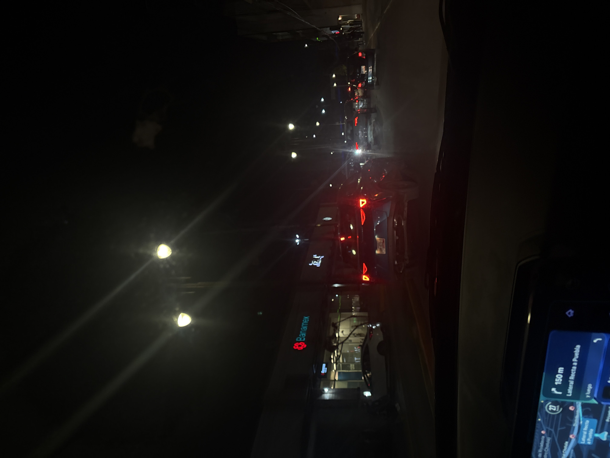The image shows a night scene from inside a car driving on a road in San Andrés Cholula, Mexico. Streetlights illuminate the road with a row of cars in front of the car from which the photo was taken. The taillights of the cars are visible. A Banamex bank branch is visible on the side of the road. A navigation system screen is visible on the right. The map shows the location is near "Lateral Recta a Puebla".