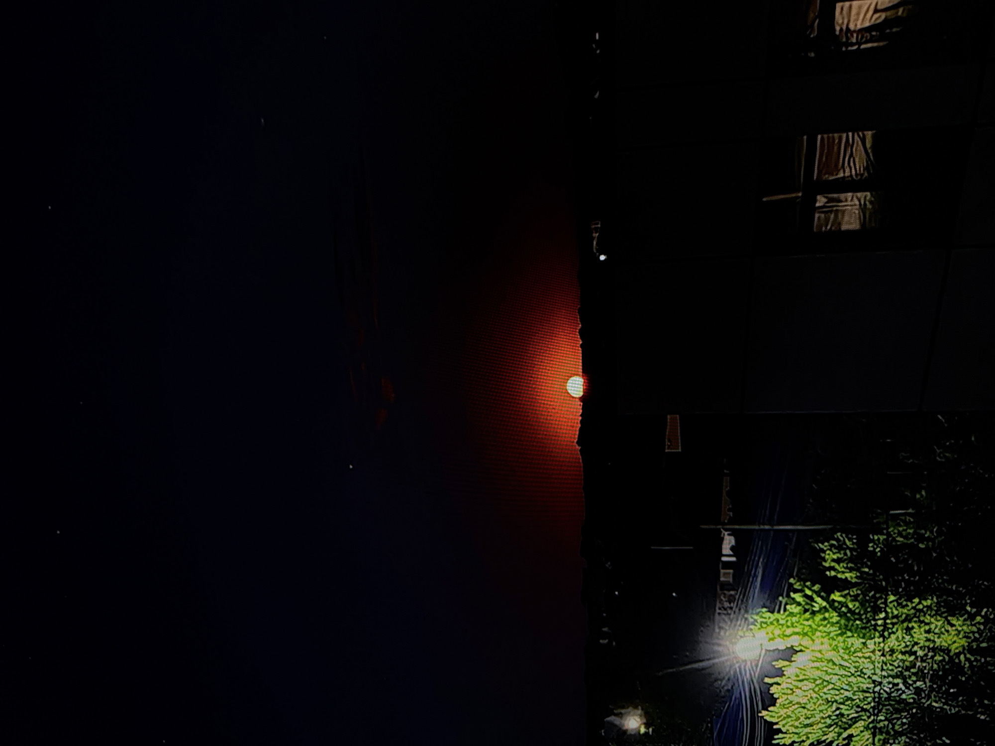 In the city of Unknown, Unknown, a sunset is captured. The sun, a vibrant orange-red orb, is setting on the horizon, partially obscured by a dark silhouette of hills or buildings. The sky above is a deep, dark blue, with a few faint stars visible. In the foreground, a bright streetlamp illuminates a lush green bush, casting a strong light on its foliage. Beyond the bush, a row of buildings and structures are dimly visible, suggesting an urban or suburban environment. The overall scene is dark and atmospheric, with the setting sun being the focal point.