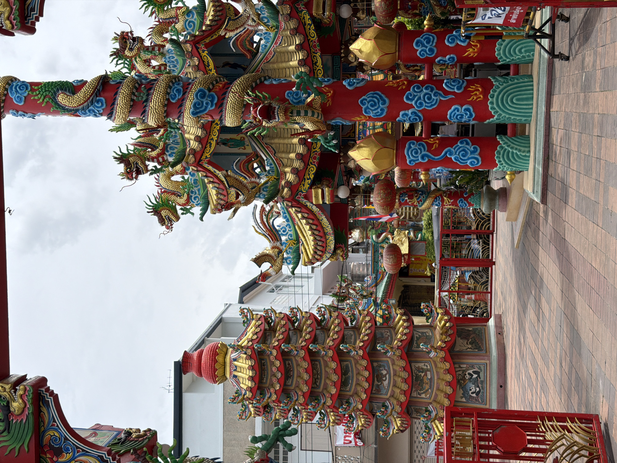 A street-level, eye-level, vertical, outdoor shot captures the ornate facade of a Chinese temple in Chiang Mai City Municipality, Thailand. The temple's architecture is predominantly red, gold, and green, with intricate dragon motifs wrapping around red pillars. A multi-tiered pagoda, also adorned with dragons, stands to the left. The foreground features a red metal gate with stylized plant designs and a brick-patterned courtyard. In the background, buildings with white and cream facades are visible, along with red lanterns hanging from the temple. The sky is overcast. No people are visible in the frame.