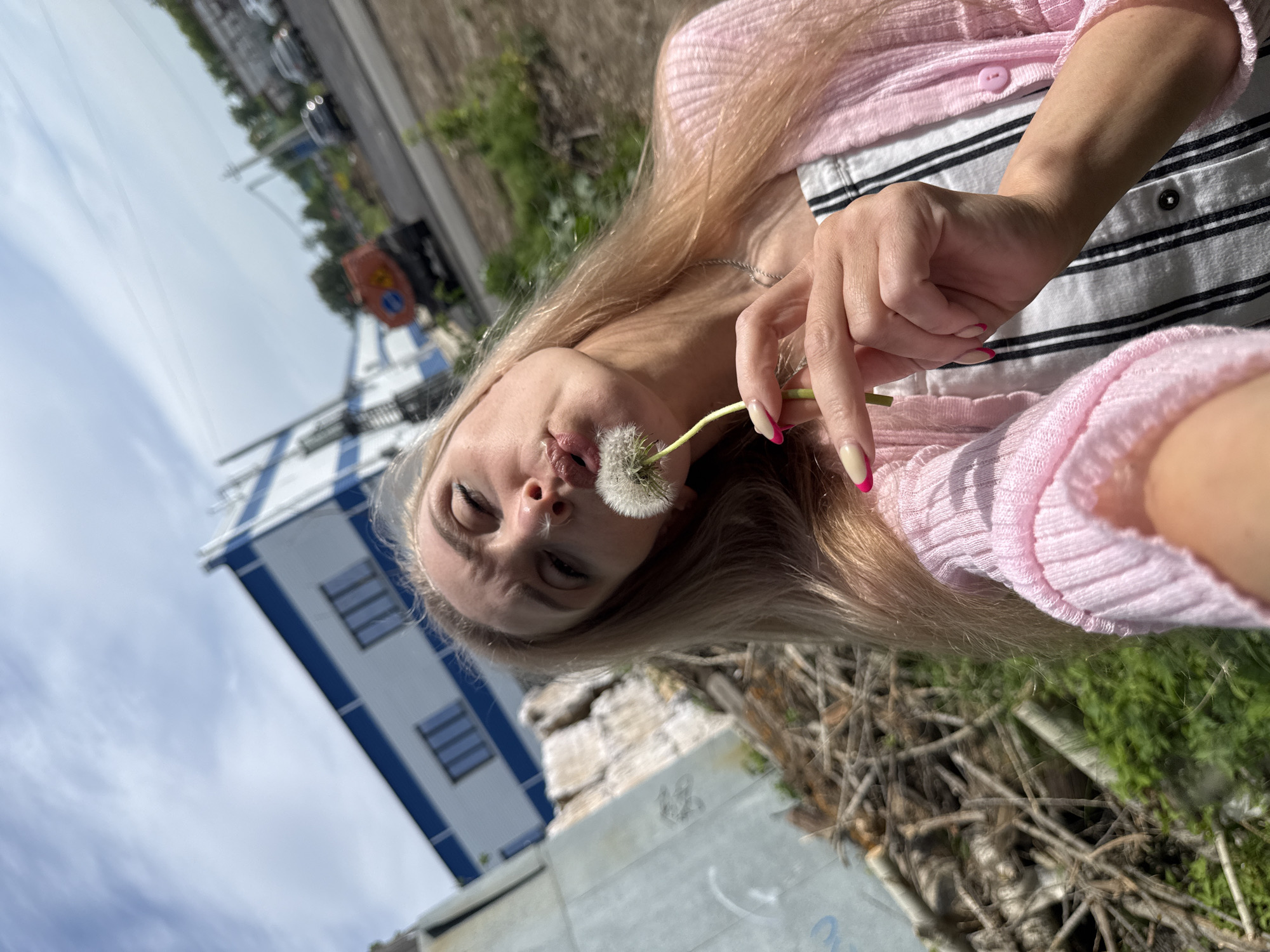 A woman with blonde hair is holding a dandelion and posing for a selfie. She is wearing a pink sweater over a striped top. The background includes a blue and white building, some greenery, concrete structures and a road with cars. The image appears to be taken outdoors during the daytime, but without further landmarks visible or specific identifying information on nearby buildings, pinpointing the location within Chaikovsky, Russia, is not possible.