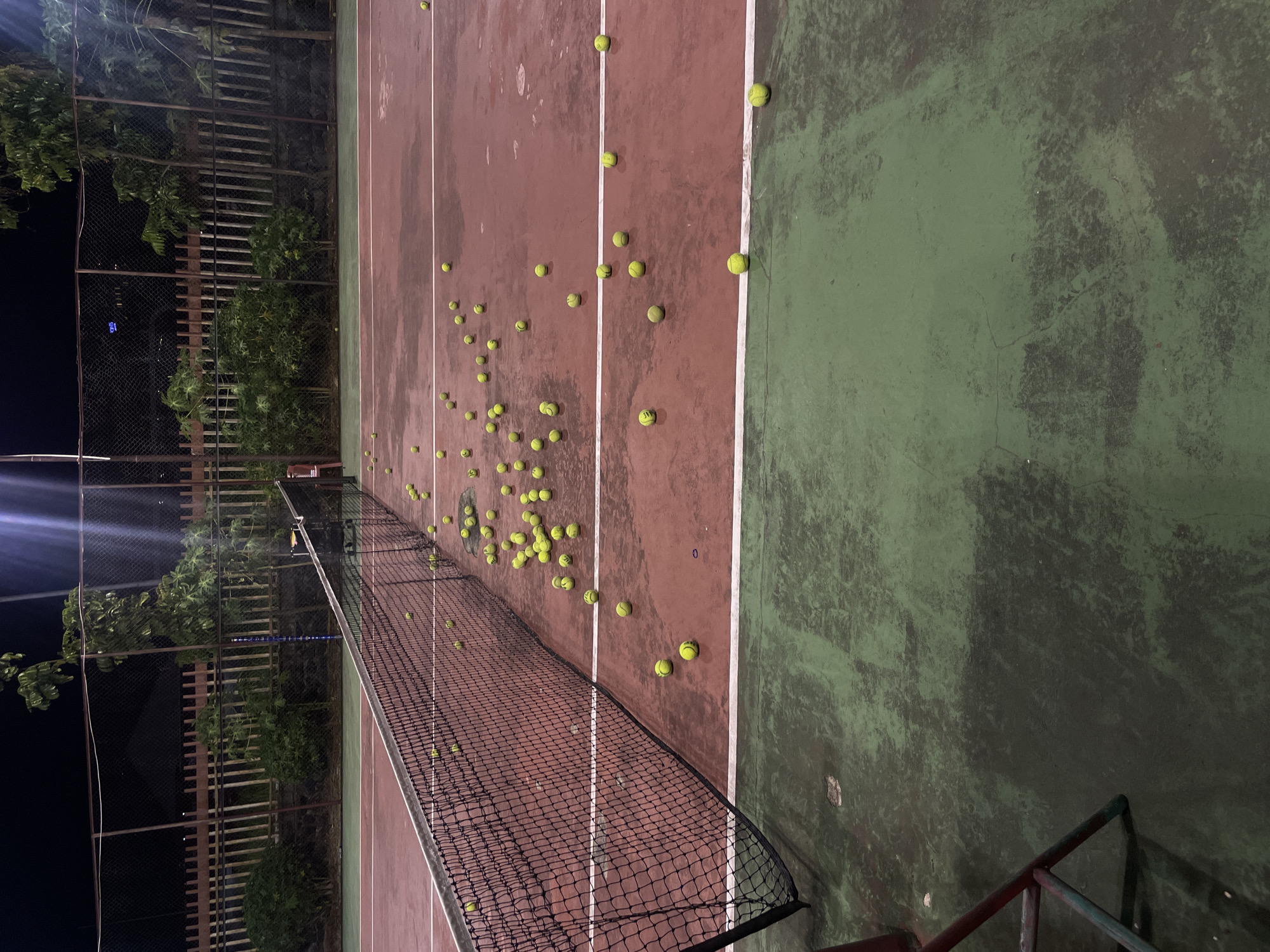 The image depicts an outdoor tennis court at night, likely located in Manado, Indonesia, based on the provided context. The court surface is a dark red with green sections. Numerous yellow tennis balls are scattered across the court, many near the net and extending into the playing area. The net is black and stretched across the width of the court. In the background, a dark fence and lush green trees are visible, with artificial lighting casting shadows and creating a somewhat moody atmosphere. There are no people visible in the scene. The court appears somewhat worn, with some patches and scuff marks visible on the surface.