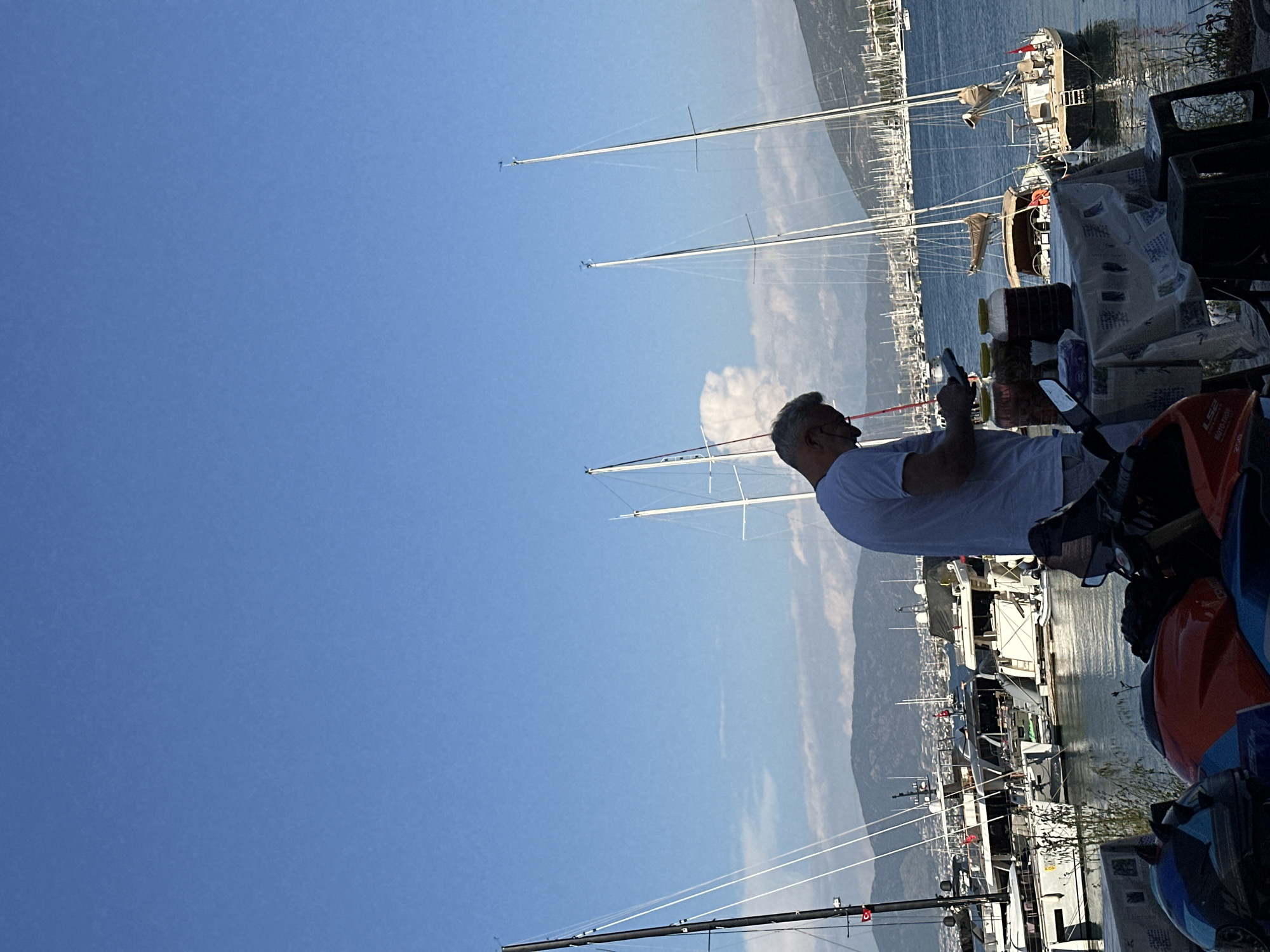 The image, taken in Fethiye, Turkey, captures a scenic marina on a bright, clear day, with the perspective rotated 90 degrees clockwise. When viewed upright, the scene depicts a man standing in the foreground on the right, facing left towards a bustling harbor. He is seen from behind, with grey hair, wearing a white short-sleeved shirt, and appears to be engrossed in looking at a small device, possibly a phone, held in his hands.
The setting is a vibrant marina filled with numerous sailboats and yachts docked in the calm blue water. Their tall masts extend upwards towards the clear, bright blue sky, with some displaying wind indicators at their peaks. In the mid-ground, a range of mountains and hills stretches across the horizon, partially obscured by a light haze, suggesting a distant landscape.
In the immediate foreground, next to the man, there are various items on what appears to be a table or surface. These include several plastic bottles with brownish liquid and patterned bags or boxes, possibly containing provisions. To the far right, a portion of an orange and blue vehicle, likely a scooter or ATV, is visible, with "LS2" and "MOTO-TRON" text partially legible on its body. A Turkish flag is visible on one of the boats in the marina, confirming the location context.
The clear sky and strong sunlight indicate it is daytime, likely late morning or early afternoon, contributing to a peaceful and leisurely atmosphere. There are no visible signs of specific events beyond the general activity of a man observing his device in a marina setting.