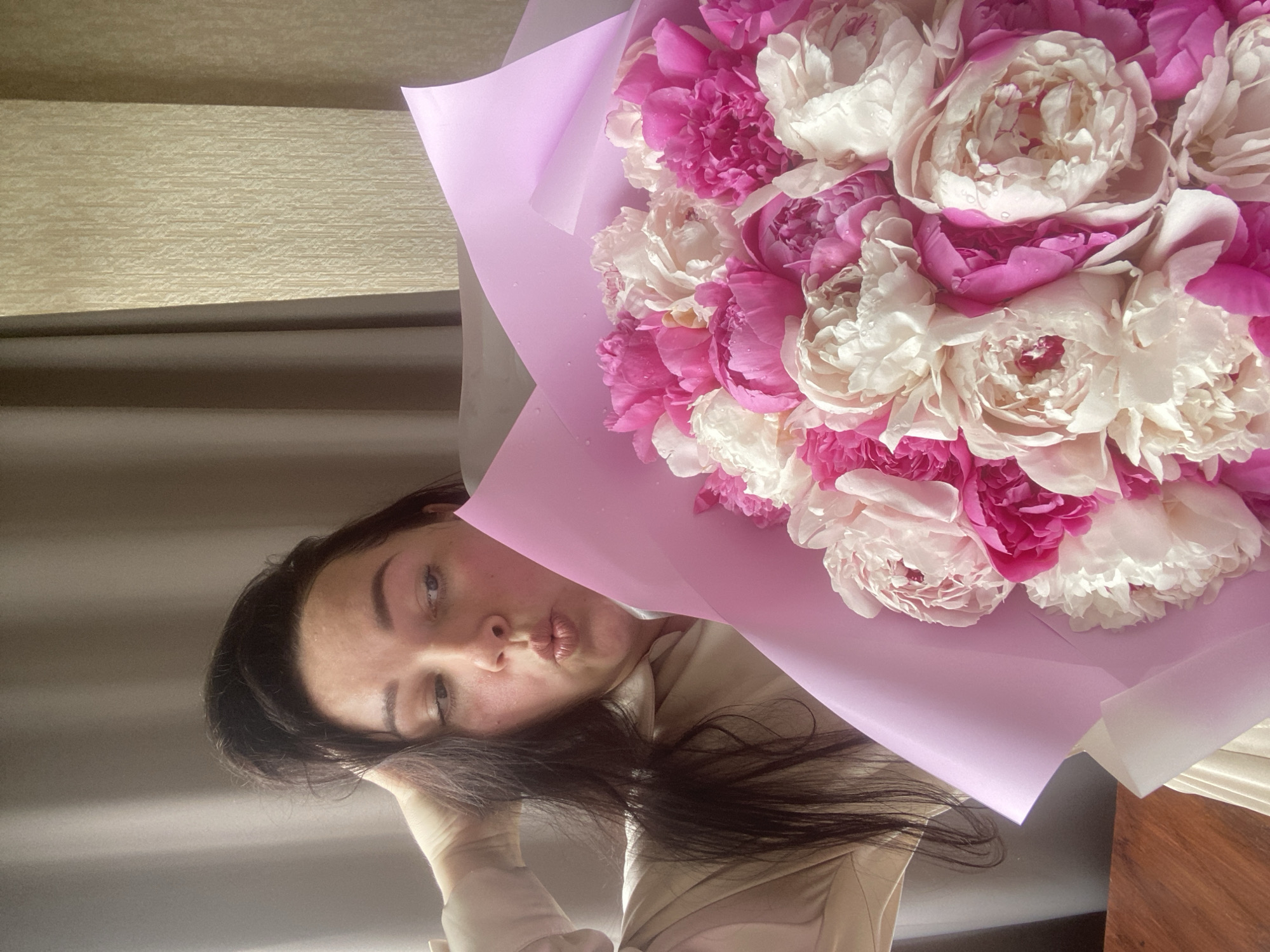 A woman with dark hair holds a large bouquet of pink and white peonies wrapped in lavender-colored paper. She is looking at the camera with a pouting expression. The background includes a textured wall and a glimpse of a window. There is no information to indicate the location is Sochi, Russia.