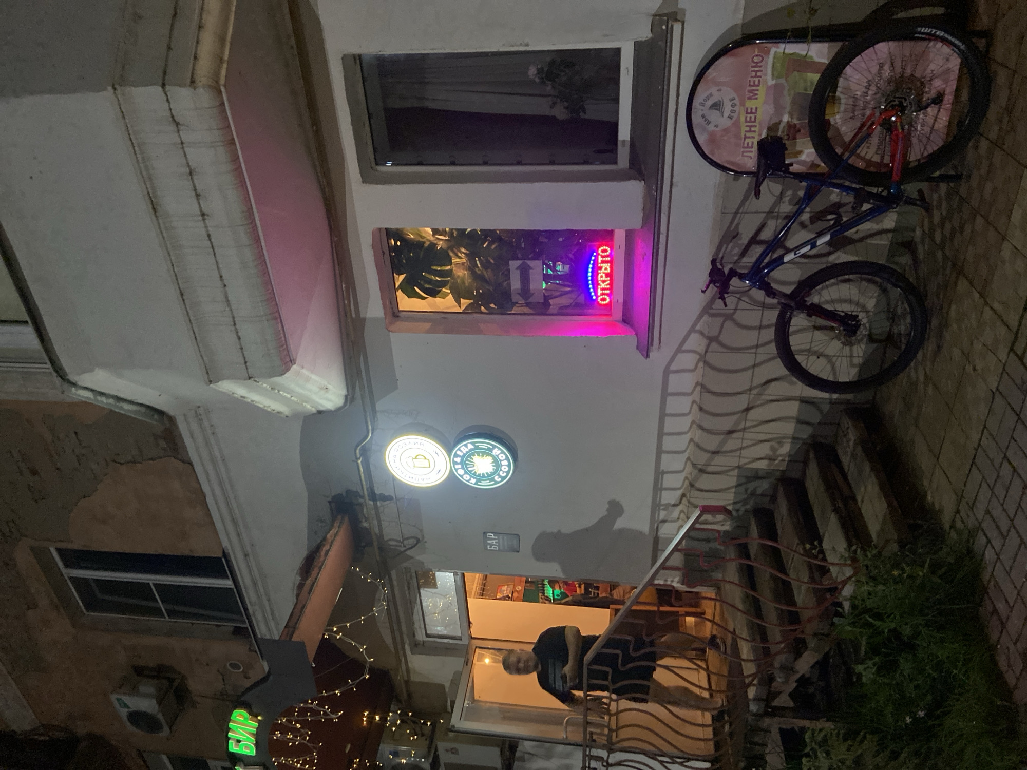 The image depicts the entrance of a bar or cafe at night in Votkinsk, Russia. A man in a dark t-shirt and shorts stands near an open doorway, holding a phone. Above the doorway, a neon sign reads "БИР" in green. Two illuminated circular signs are mounted on the wall to the right of the entrance: one with a beer mug and text, the other with a sunburst graphic and text. A smaller sign that reads "БАР" is also visible on the wall. A neon sign in a window displays the word "ОТКРЫТО" (Open) in pink. The window to the right shows a curtain and a potted plant. A bicycle with a blue frame and red accents is parked next to a freestanding sign advertising a "ЛЕТНЕЕ МЕНЮ" (Summer Menu). The building appears to be made of stucco with some signs of wear. The ground is paved with brick and cobblestones, and there is a set of wooden stairs with a decorative metal railing leading to the entrance. Lights are strung across the awning above the entrance, and interior lights illuminate the scene.